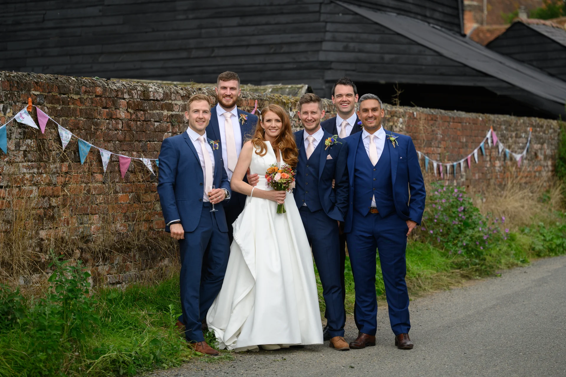 The radiant bride holding a colourful bouquet stands proudly with her groomsmen in matching navy suits alongside a rustic brick wall decorated with bunting.