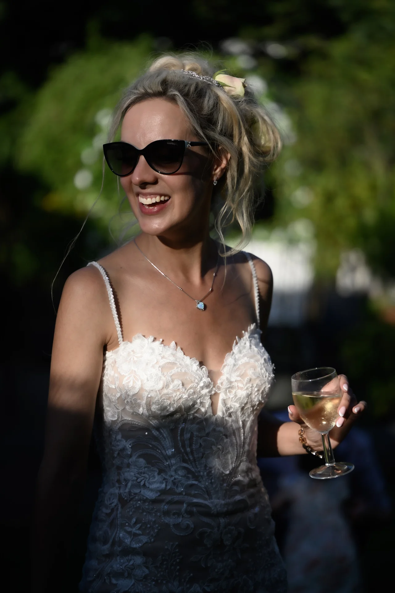 A smiling bride wearing sunglasses and a lace wedding dress holds a glass of champagne during an outdoor drinks reception.