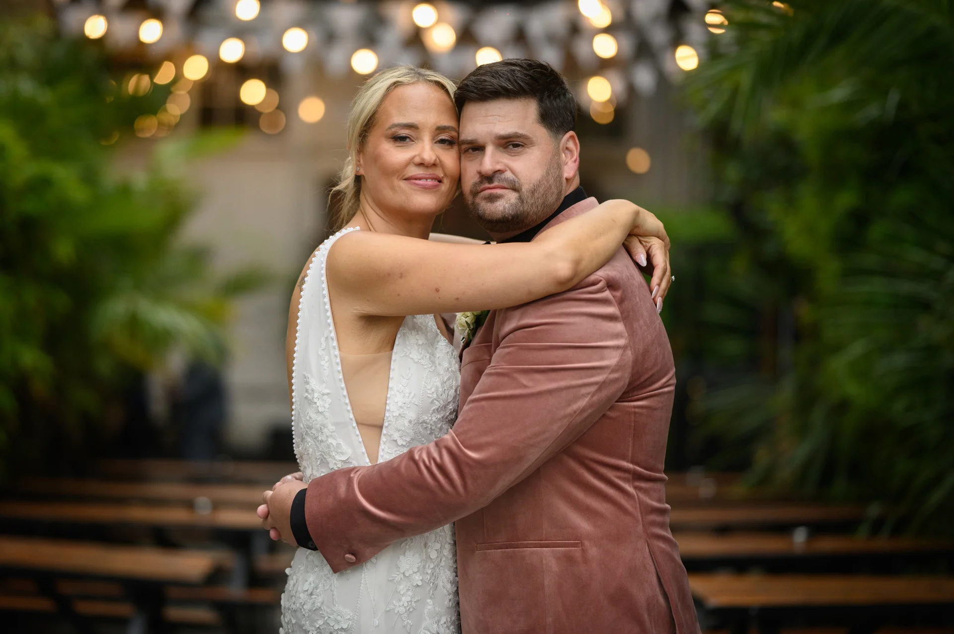 A bride and groom share a tender embrace outdoors under glowing fairy lights, with the groom wearing a velvet jacket and the bride in a lace dress.