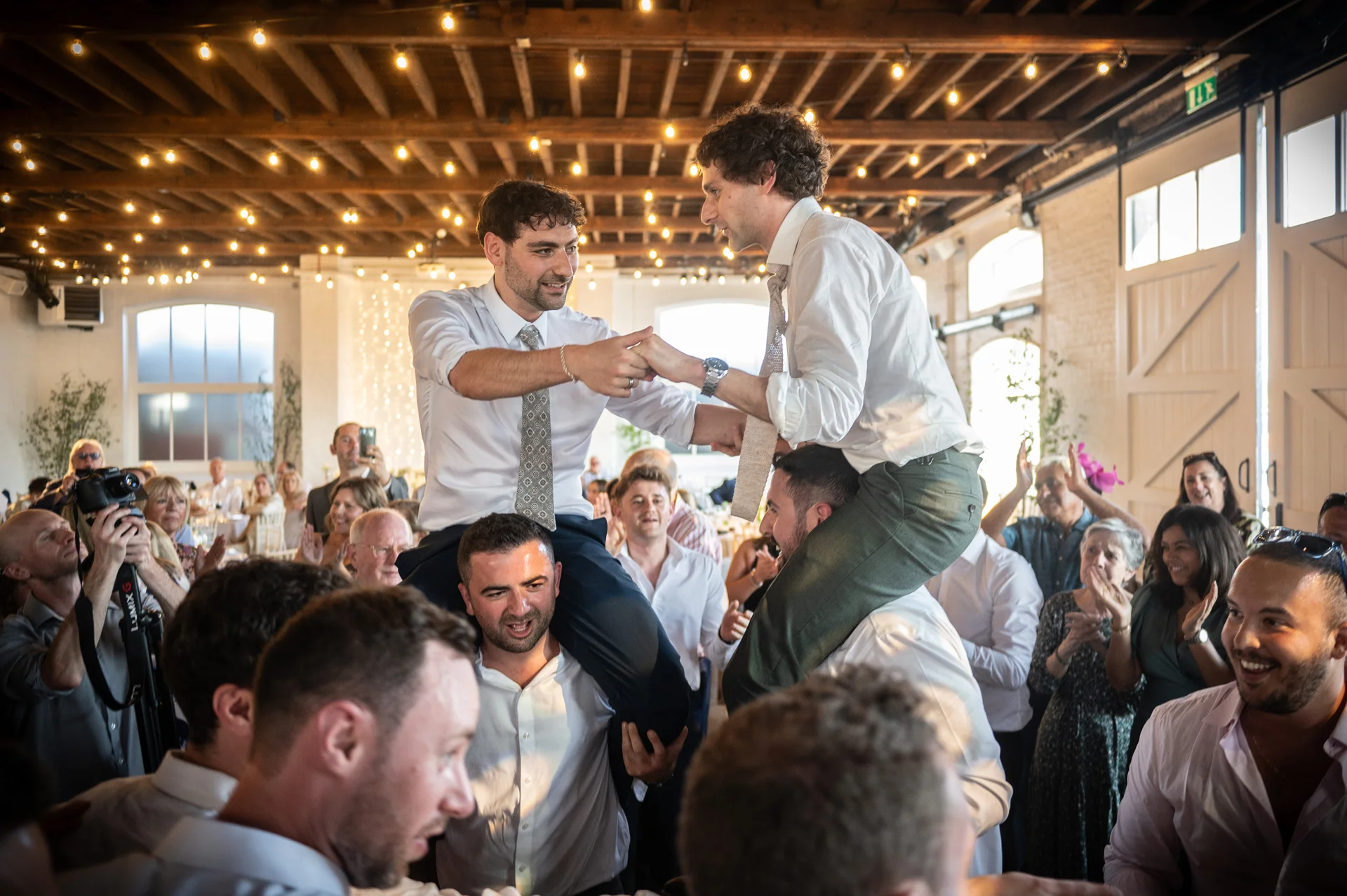 Two grooms are lifted onto the shoulders of guests inside a rustic warehouse venue illuminated by strings of warm fairy lights.
