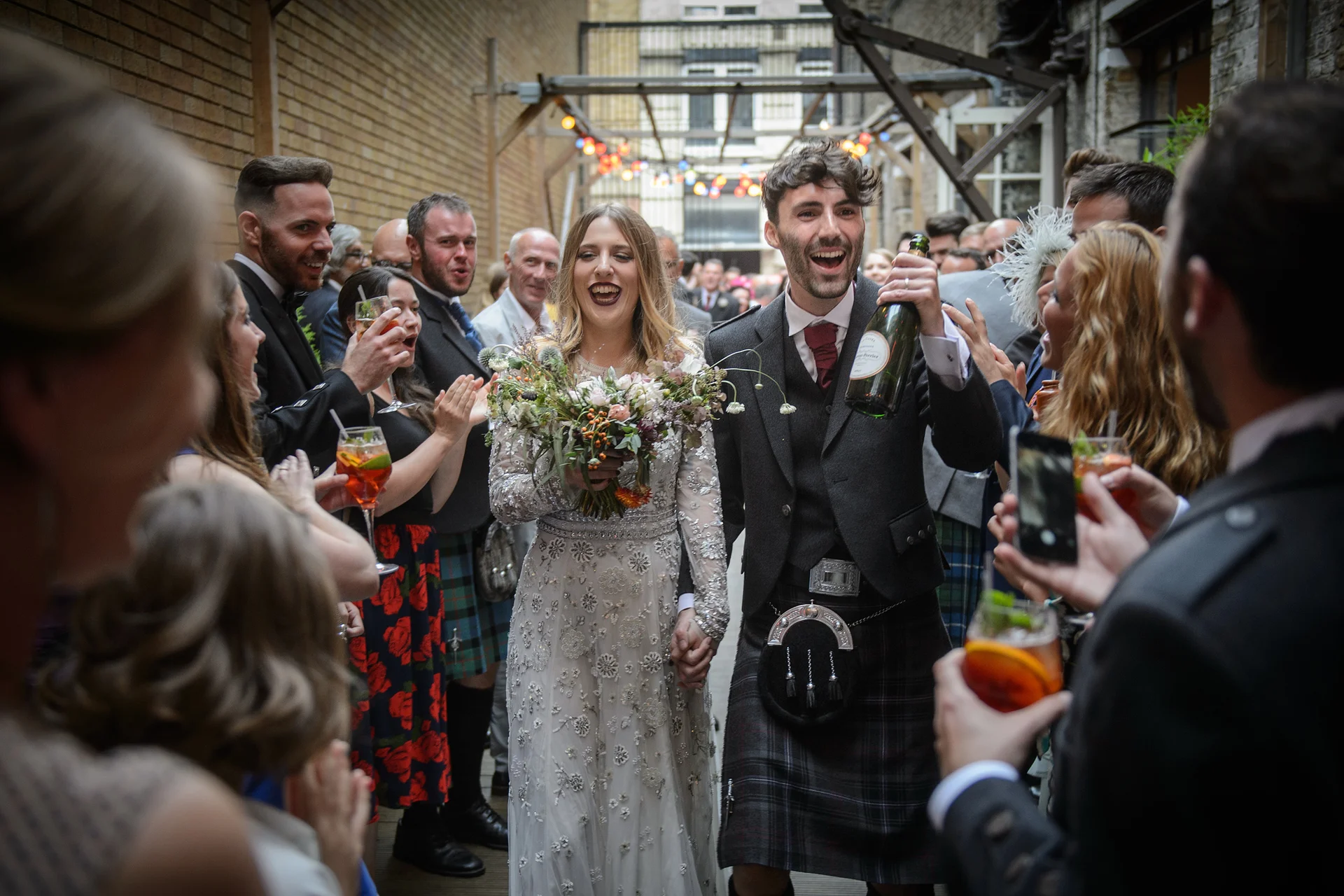 A joyful newlywed couple walks through an urban courtyard flanked by cheering guests holding drinks while the groom raises a bottle of champagne in celebration.