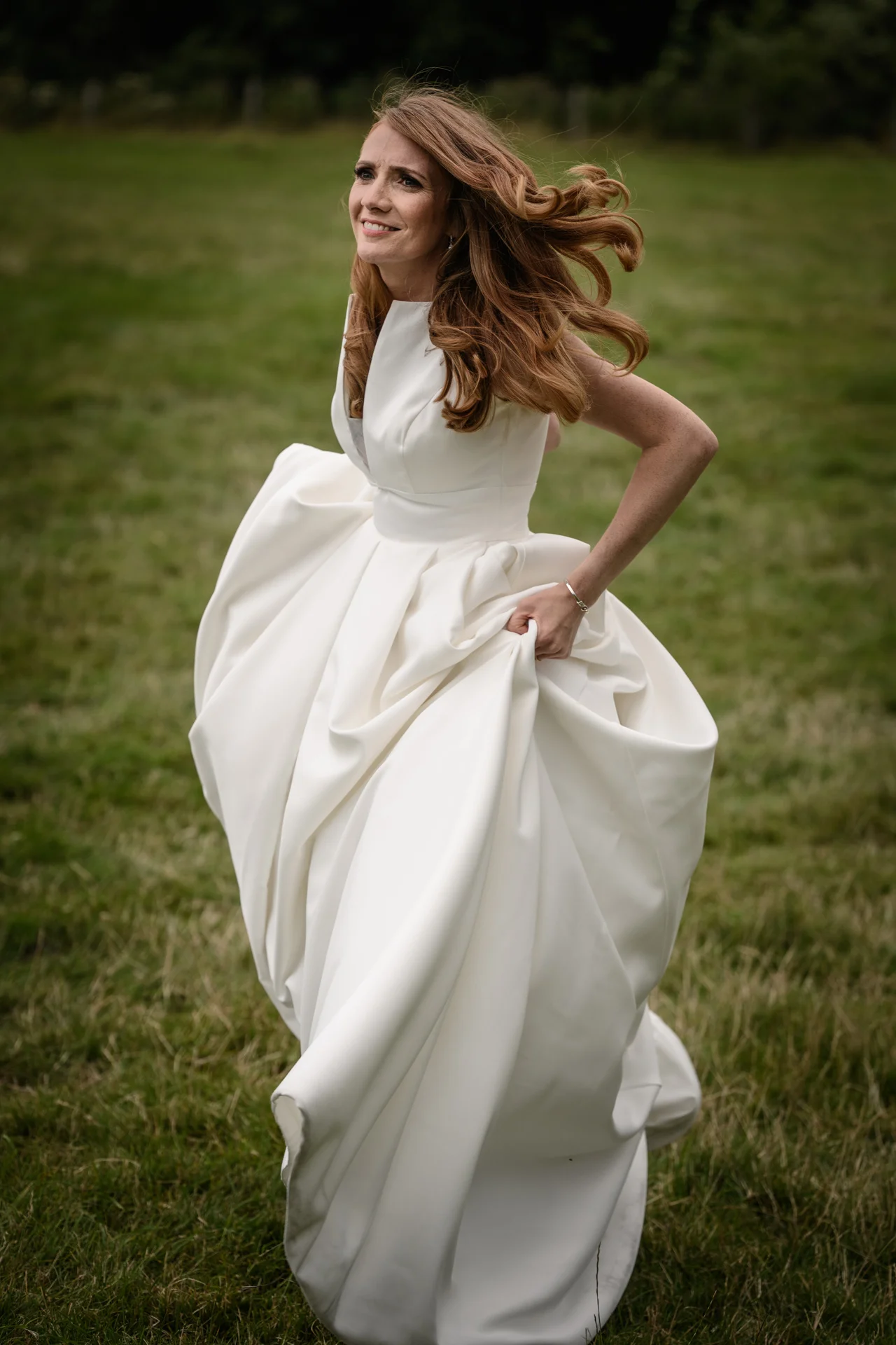 A joyful bride with long red hair runs through a grassy field, holding up the voluminous skirt of her white satin wedding gown.