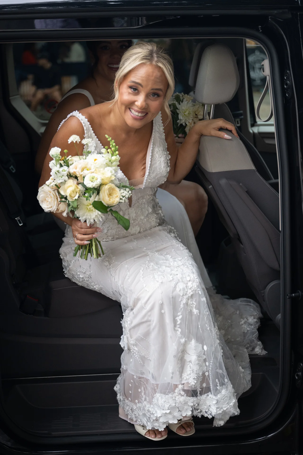 A smiling bride wearing an embroidered floral gown and holding a white bouquet steps out of a black wedding car.