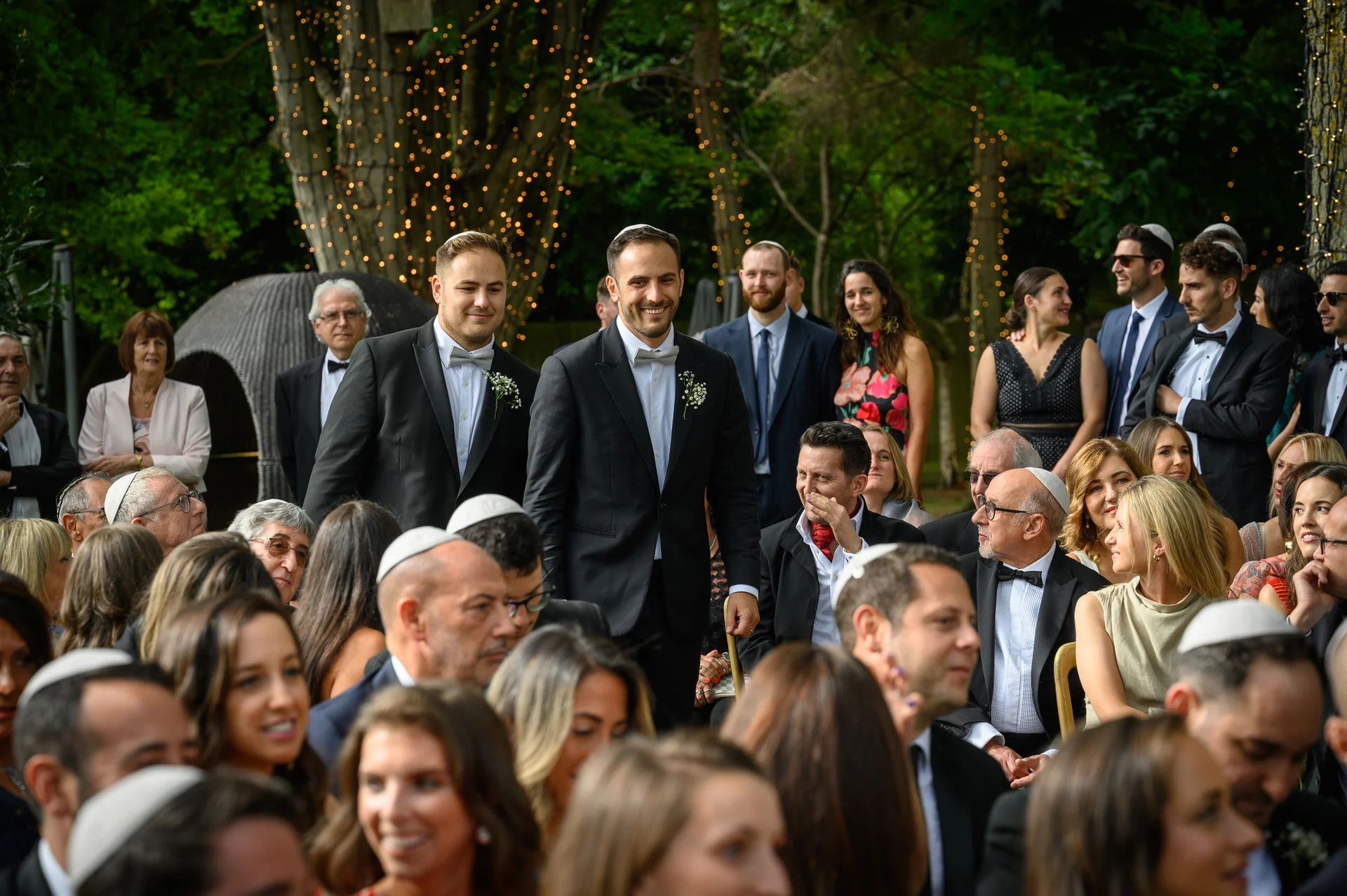 Two grooms in matching black tuxedos and bow ties smile happily together while walking through an outdoor wedding ceremony surrounded by seated guests and trees adorned with string lights.