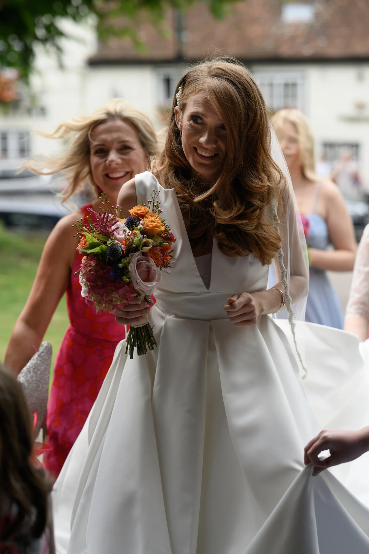 A joyful bride smiles as she holds a colourful hand-tied bouquet, wearing a satin v-neck gown and veil while arriving outdoors.