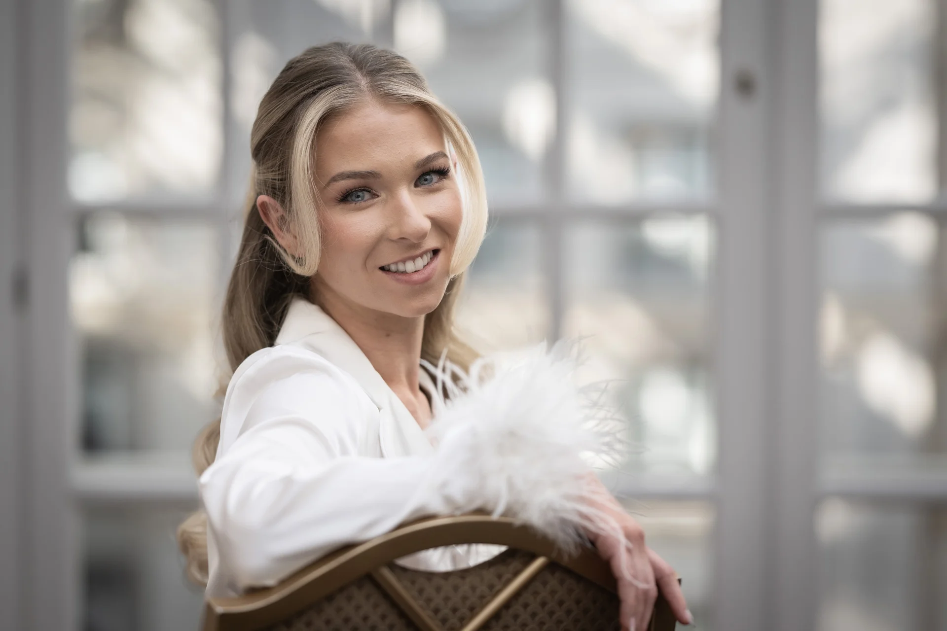 A smiling bride wearing a white outfit with feathered cuffs looks over her shoulder while posing backward on a chair in front of a window.