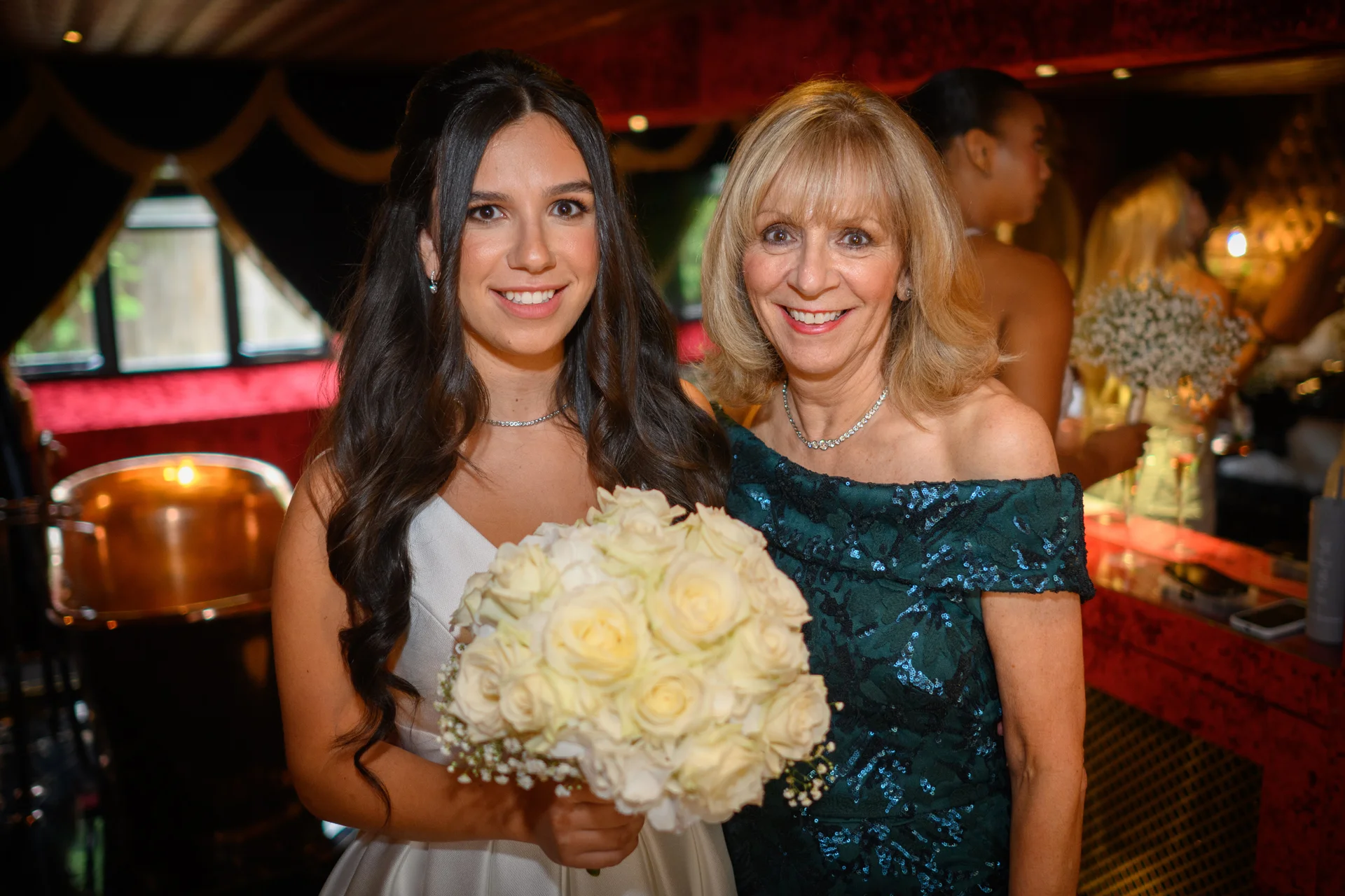 A smiling bride holding a white rose bouquet poses alongside her mother wearing an off-the-shoulder green sequin dress at an evening indoor wedding reception.