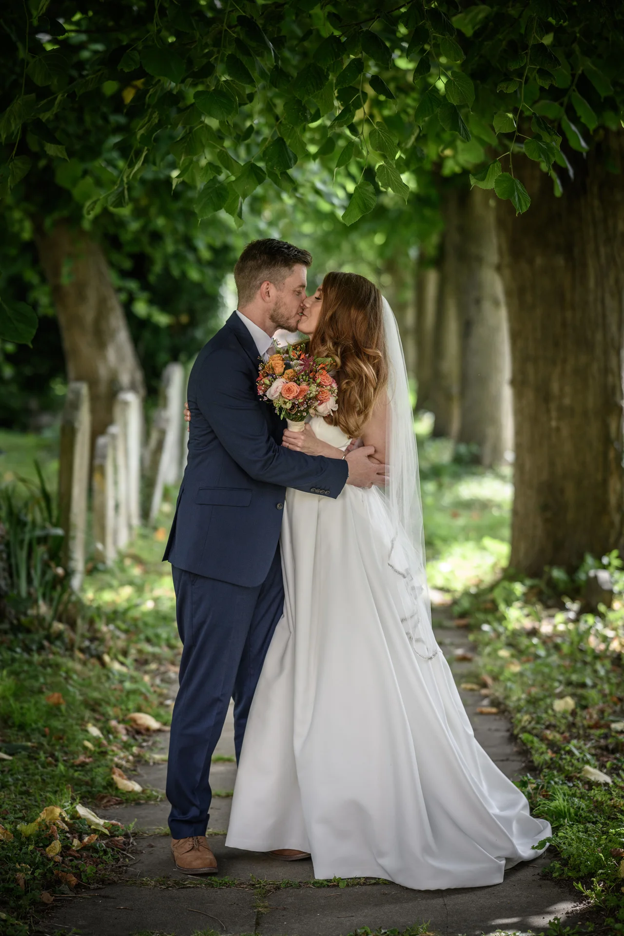 A newlywed couple shares a tender kiss along a tree-lined pathway framed by overhanging foliage during their outdoor wedding portrait session.