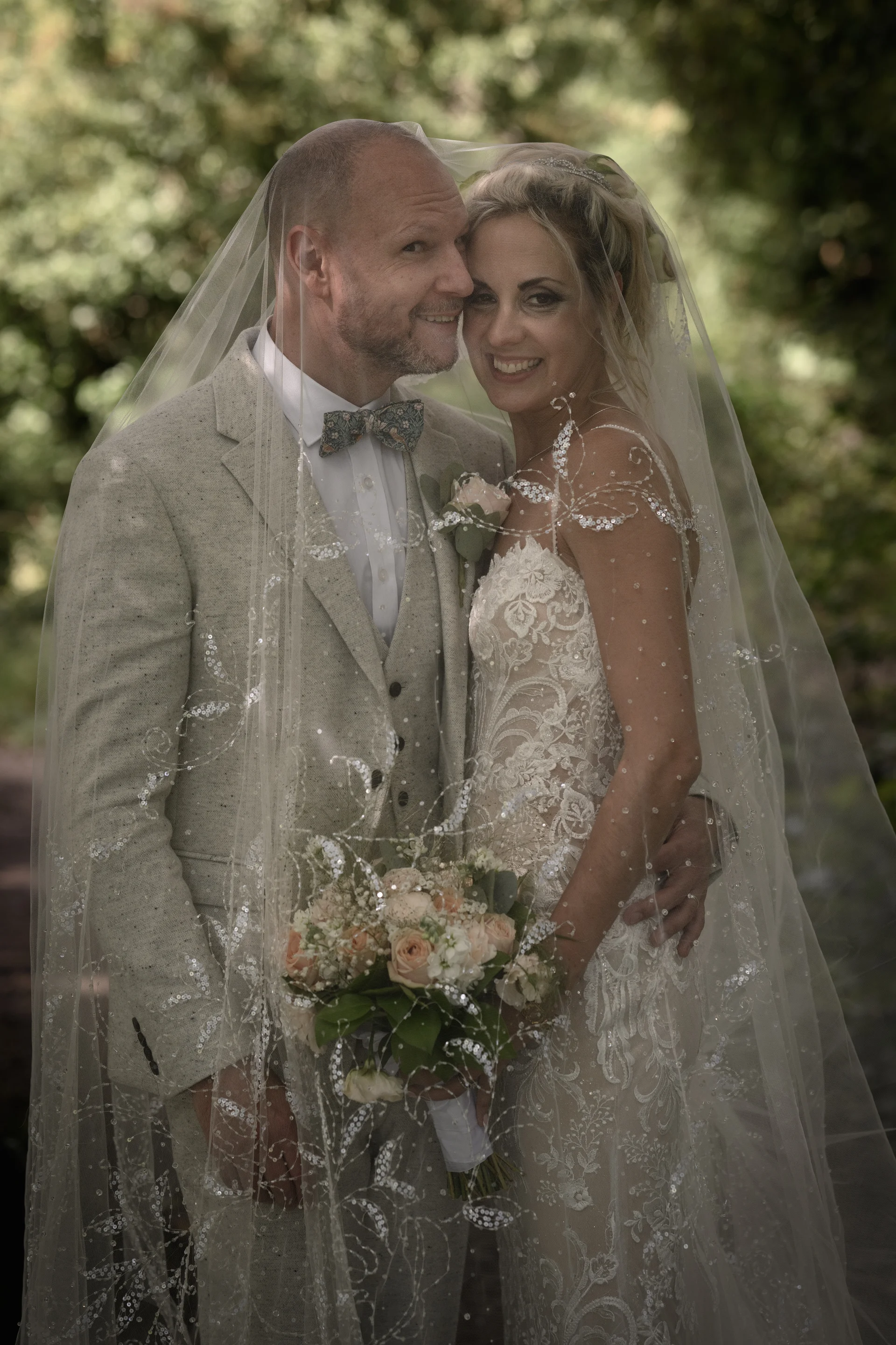 A smiling bride and groom stand closely together in a wooded setting, sharing a tender moment beneath an intricately embellished veil while holding a floral bouquet.