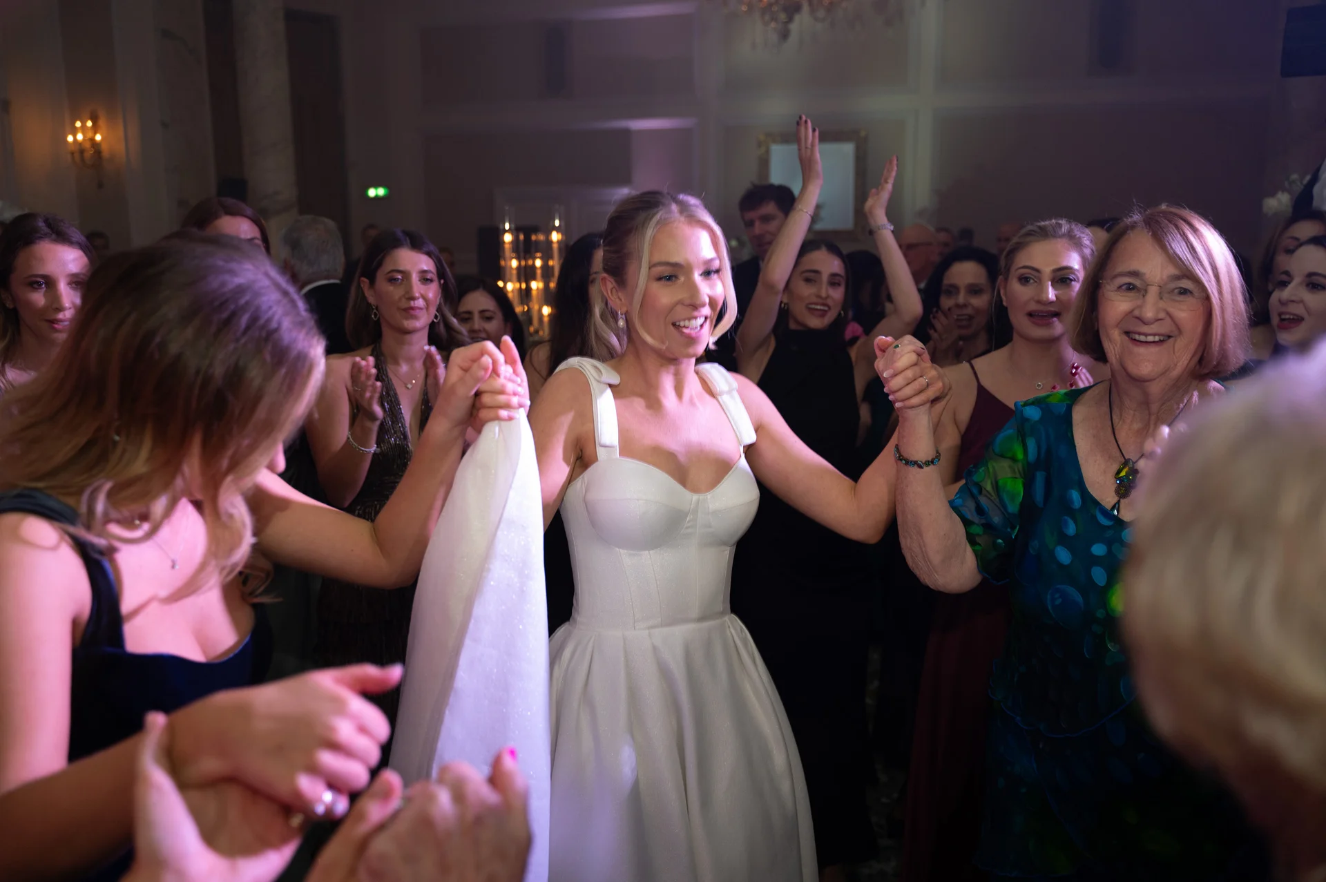 A joyful bride in a structured white gown smiles brightly while dancing in a circle with her wedding guests during the evening reception.