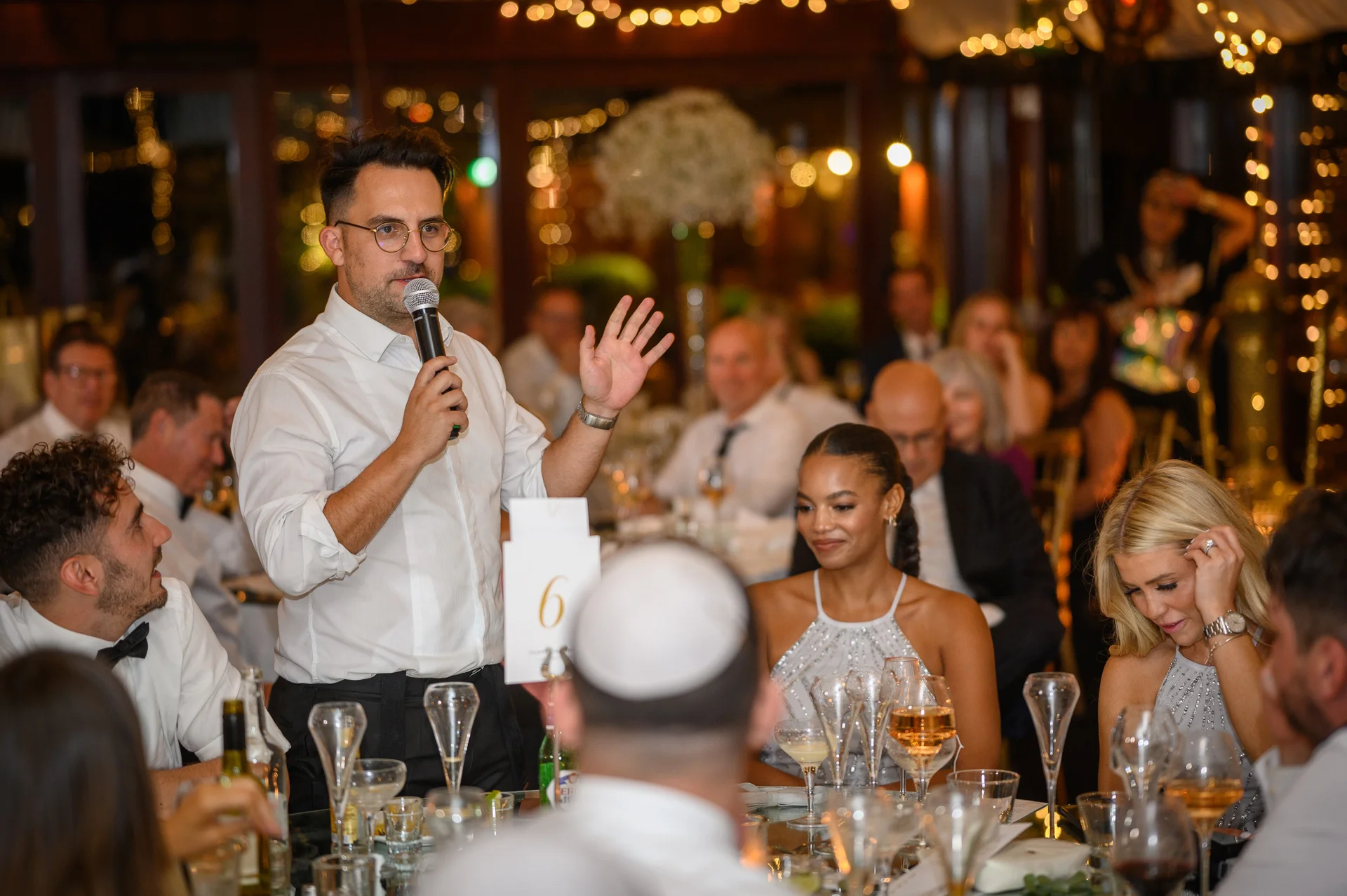 A groom delivers a speech into a microphone during a wedding reception, gesturing with his hand while seated guests watch and laugh at tables illuminated by fairy lights.
