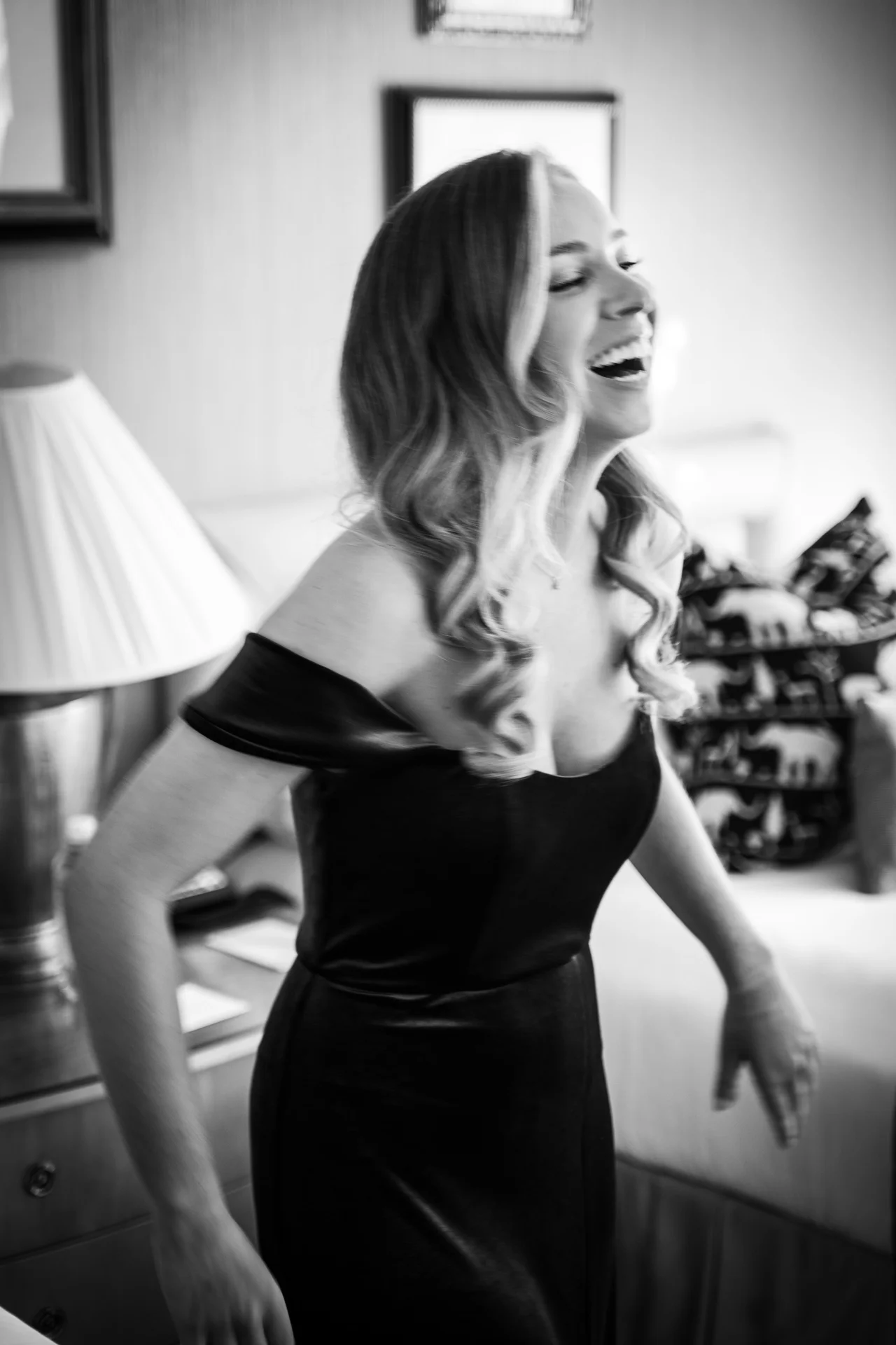 A joyful bridesmaid wearing an off-the-shoulder black dress laughs heartily indoors during wedding morning preparations.