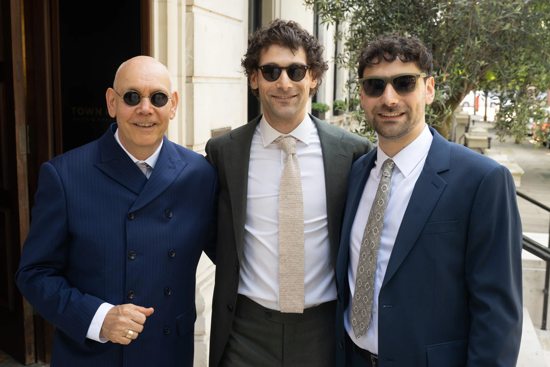 Three smiling male wedding guests pose together outdoors wearing smart suits and stylish sunglasses outside a hotel venue.