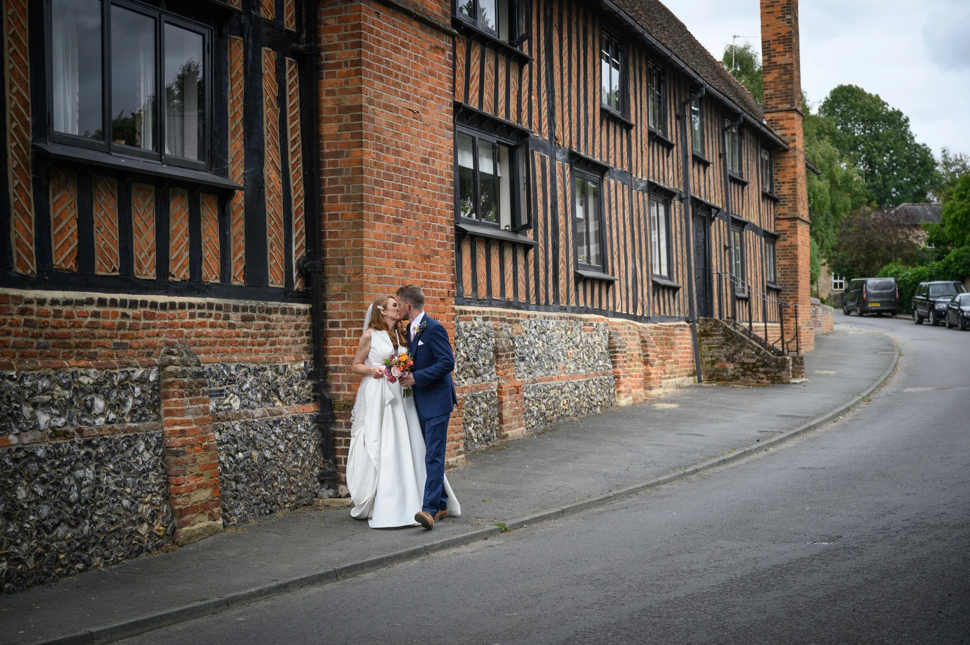 A newlywed couple shares a tender kiss while standing on a pavement alongside a historic timber framed and flint building.
