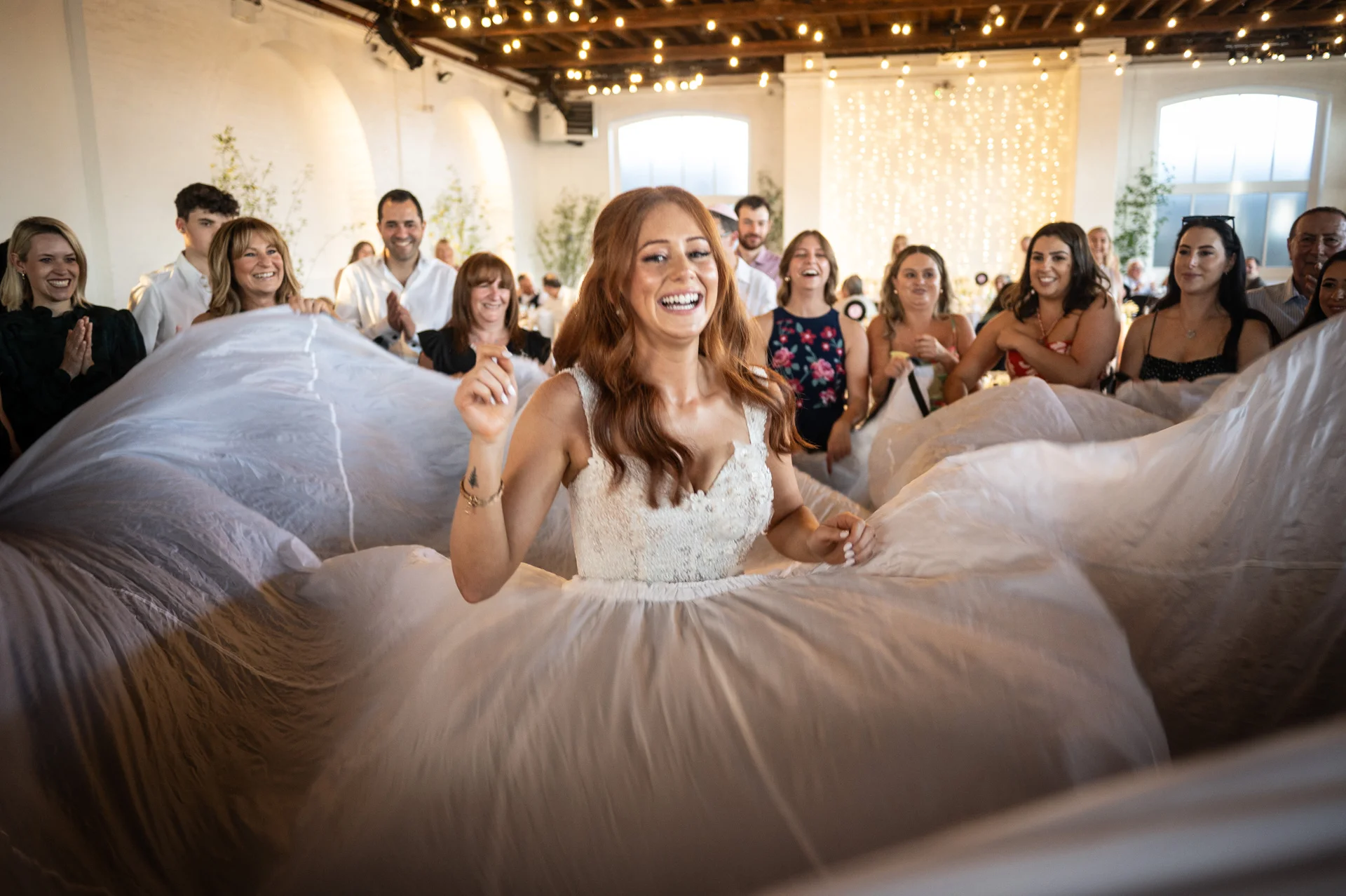 A joyful red-haired bride laughs excitedly as she wears a dramatic tulle skirt surrounded by cheering wedding guests inside an industrial warehouse venue.