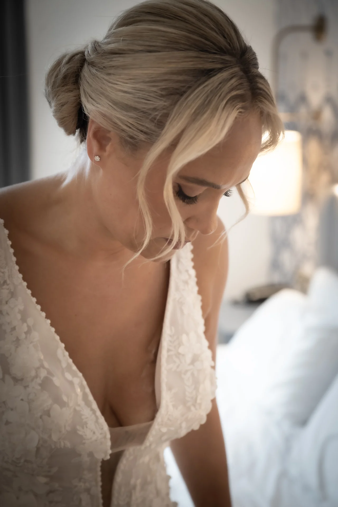 A stunning bride looking down thoughtfully while getting ready, wearing a lace v-neck dress and a soft updo with face-framing tendrils.