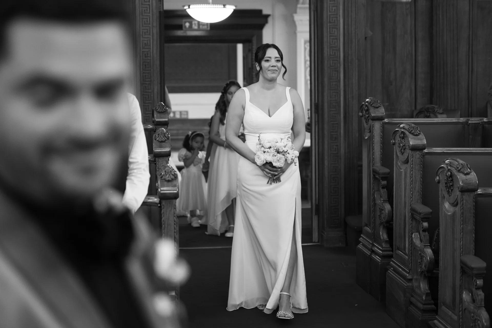 The bride smiles as she walks down the aisle holding a bouquet, viewed past the out-of-focus groom waiting at the front of the venue.