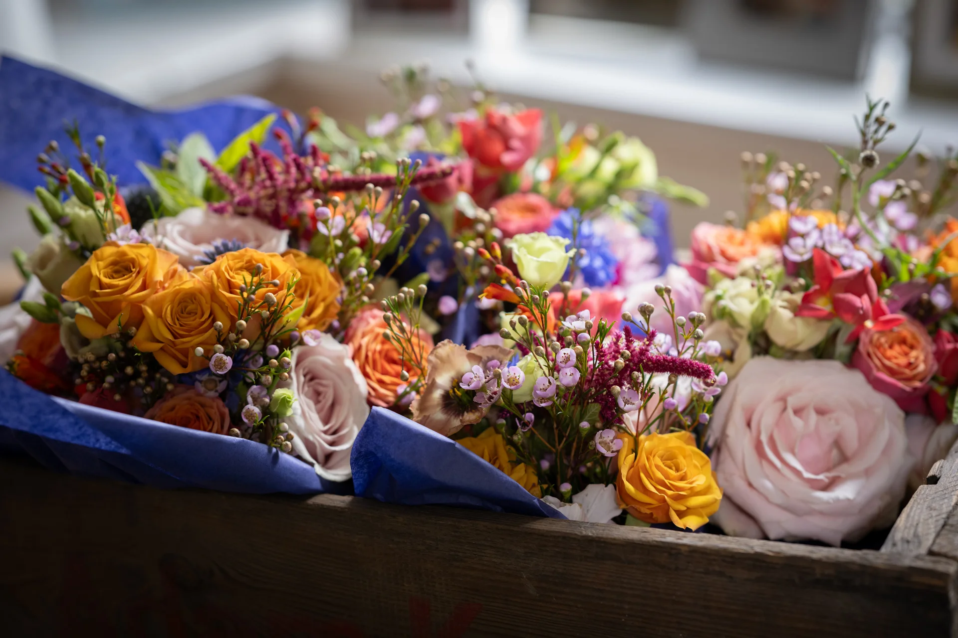 A rustic wooden crate filled with vibrant, hand-tied wedding bouquets featuring colourful roses and delicate pink waxflower.
