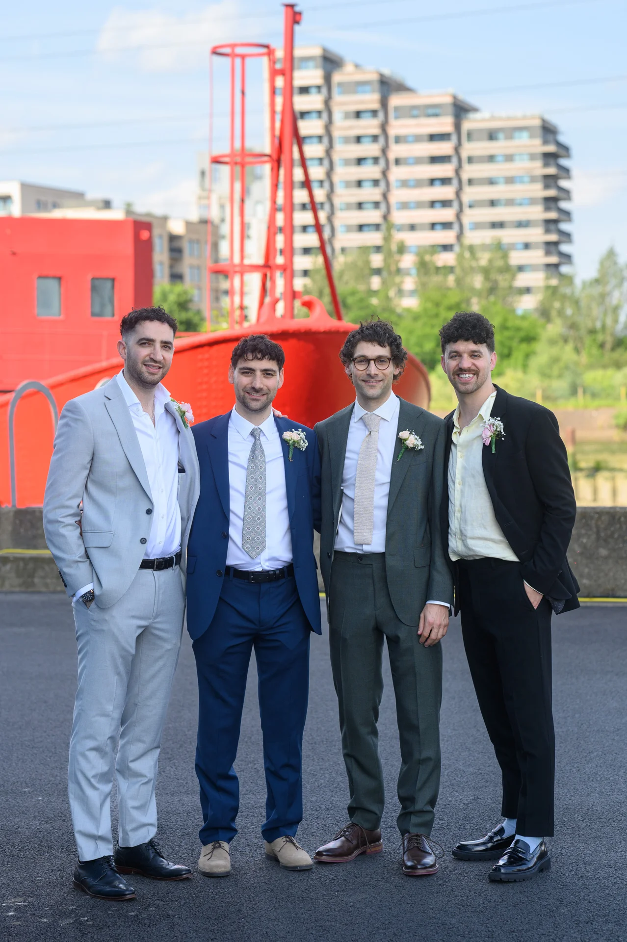 Four groomsmen wearing mismatched suits and buttonholes pose together outdoors in front of a striking red industrial structure and modern city buildings.
