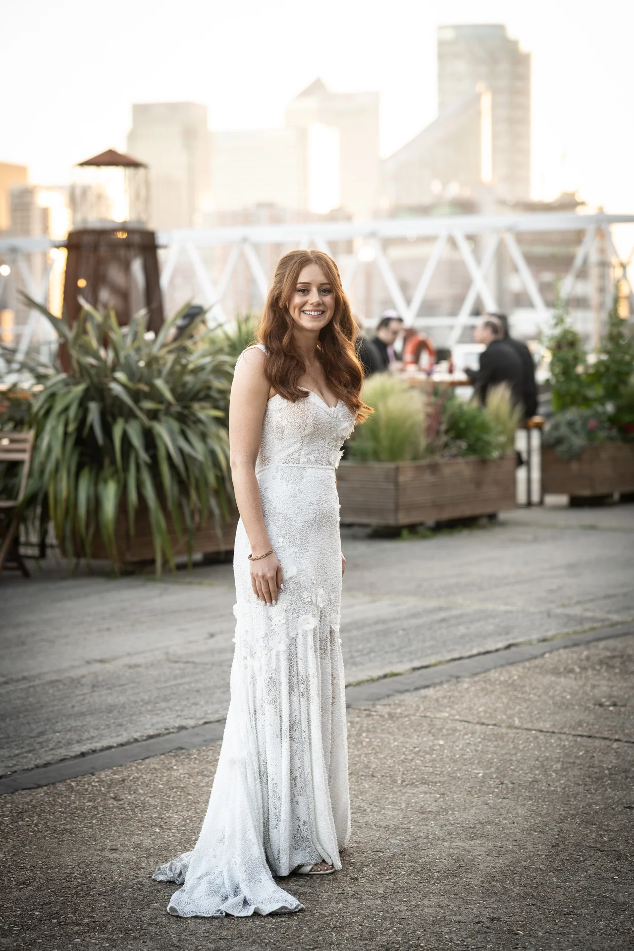 A stunning solo bride smiling in a beaded strapless column wedding dress during golden hour with a city skyline backdrop.