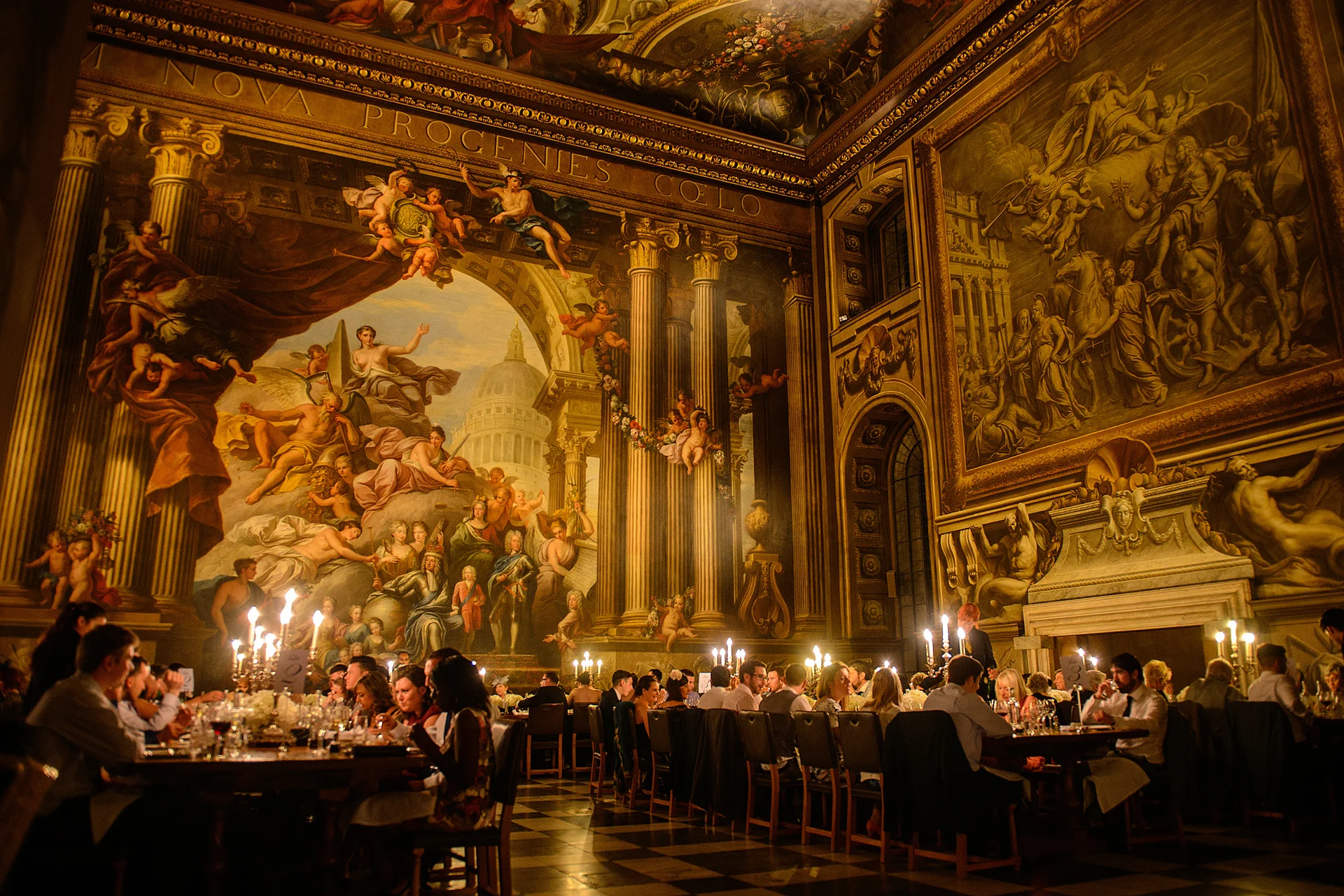 Guests enjoy a candlelit wedding breakfast inside an opulent historic hall featuring dramatic wall murals and soaring gilded architecture.
