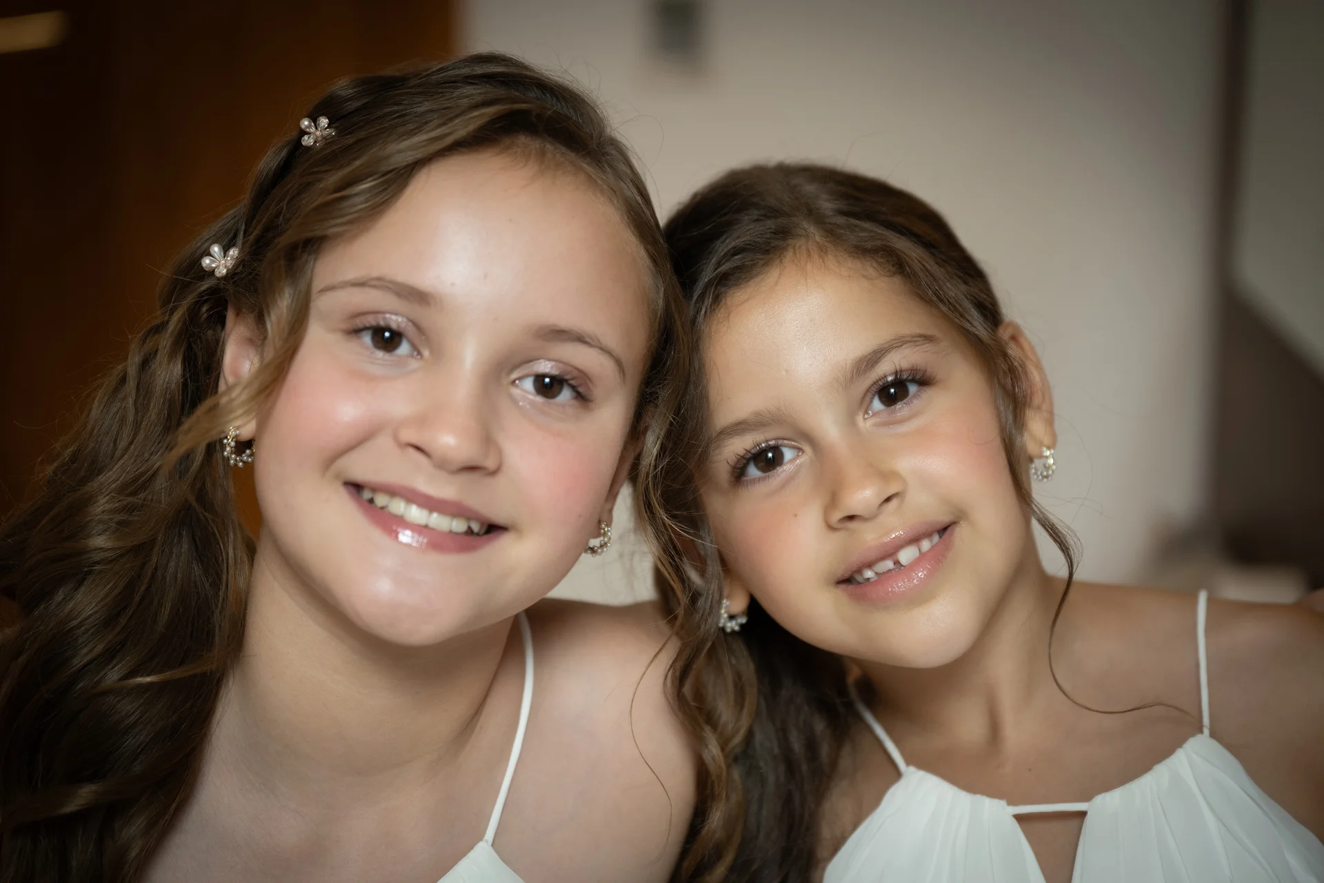 Two young junior bridesmaids with smiling faces pose closely together wearing matching white dresses and delicate hair accessories.