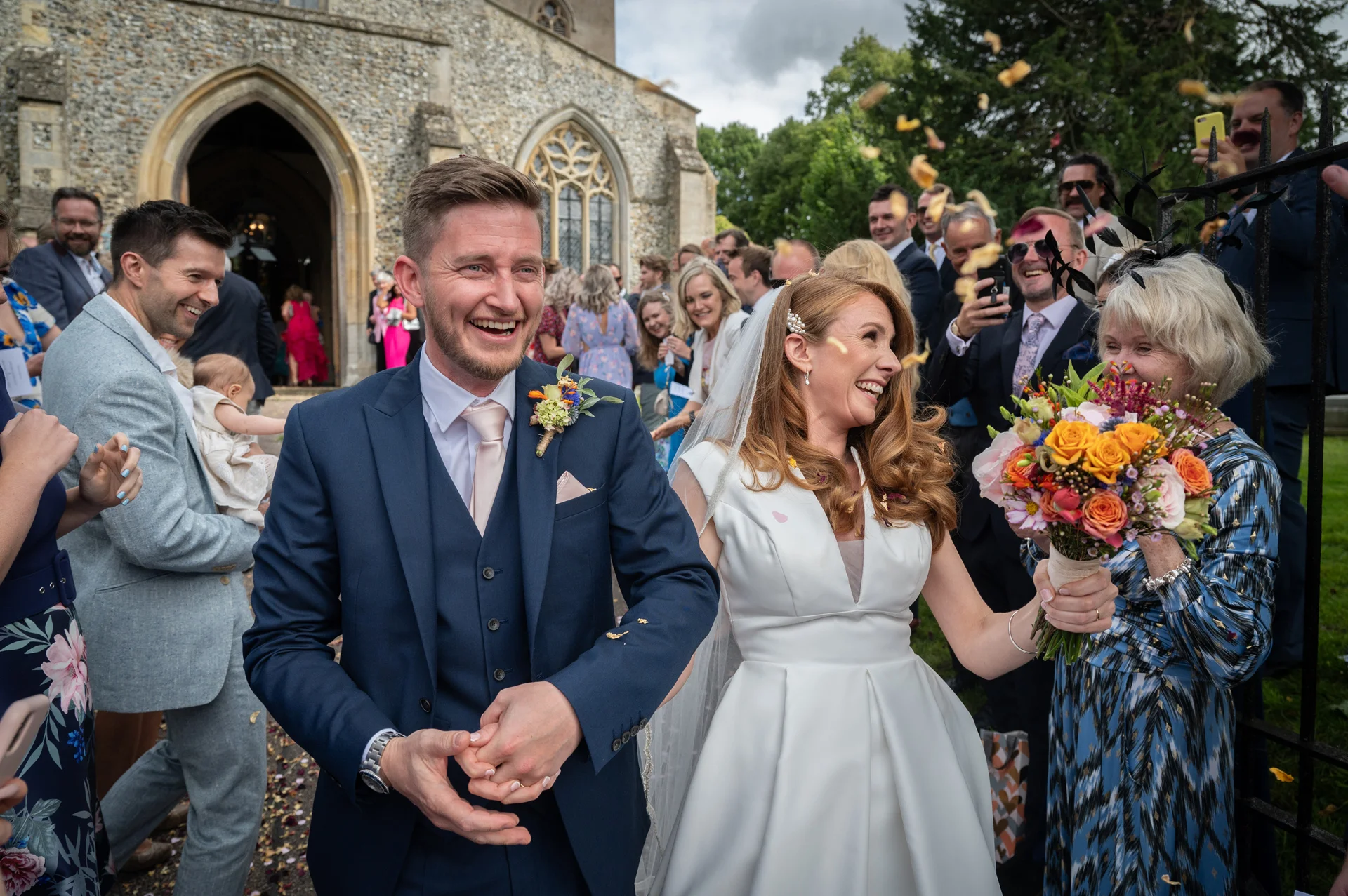 A joyful newlywed couple walks hand in hand out of a stone church while guests shower them with confetti.
