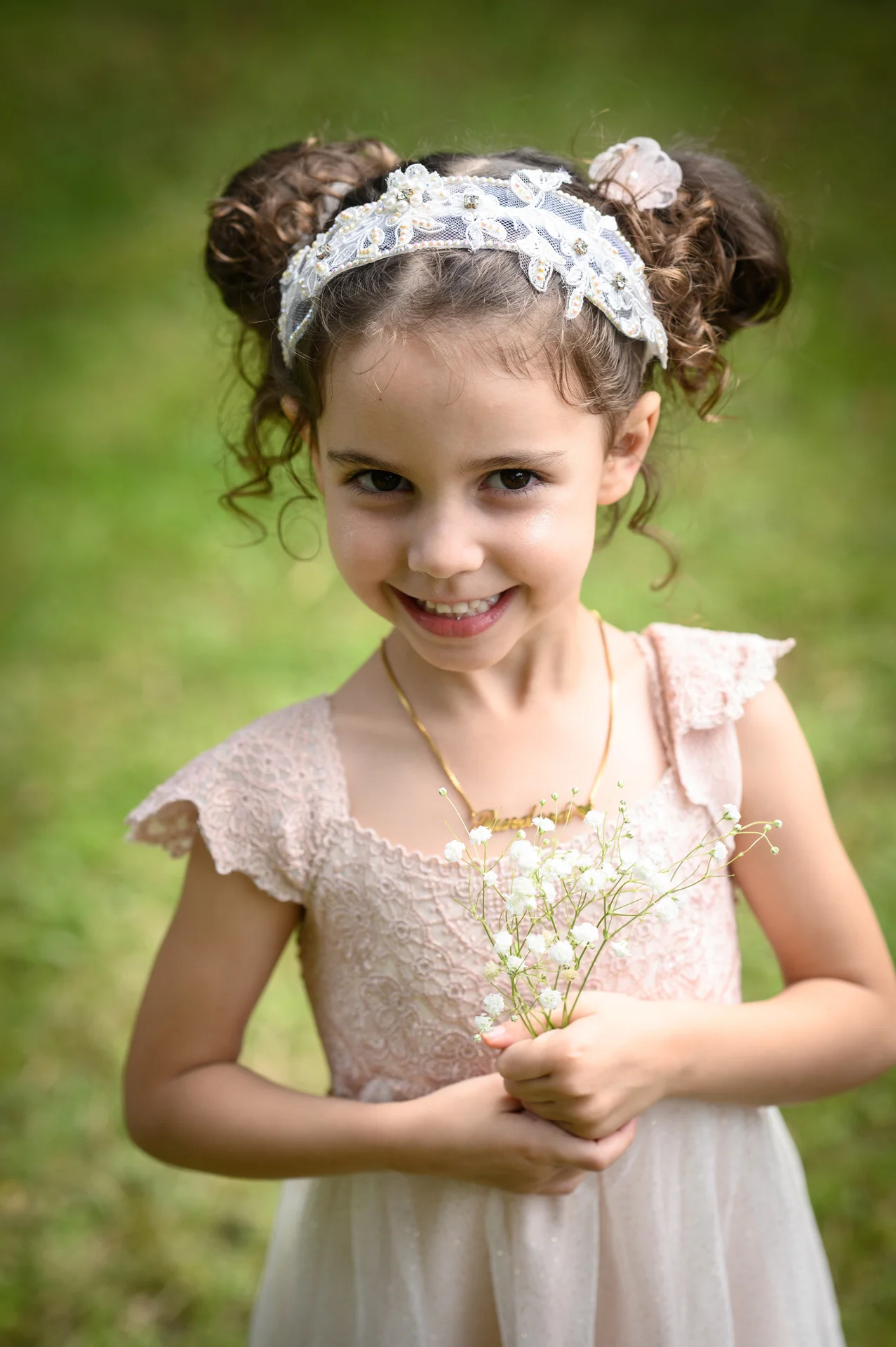 A young flower girl with curly hair styled in double buns and wearing an embellished lace headband smiles while holding a delicate bunch of baby's breath outdoors.