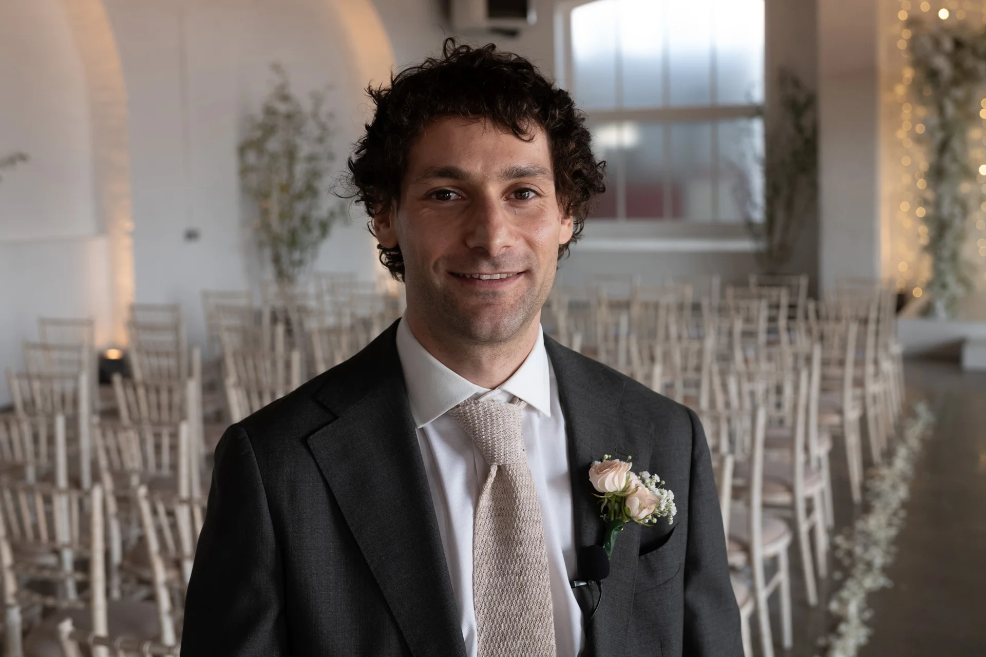 A smiling groom with dark curly hair wearing a charcoal suit and a textured neutral tie stands in a light-filled ceremony room with rows of chairs.
