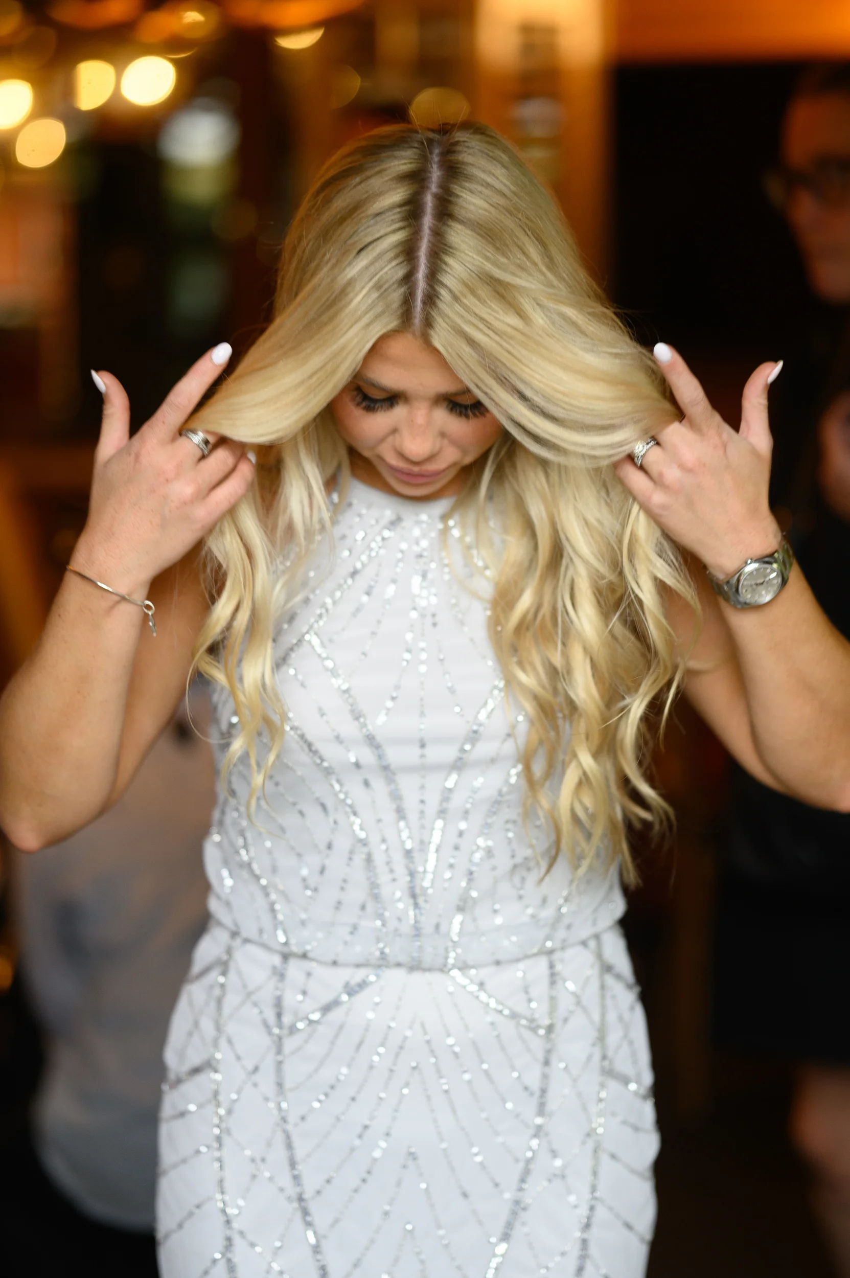 A blonde bride wearing a crystal-embellished high-neck gown gently adjusts her long, wavy hair during morning preparations.