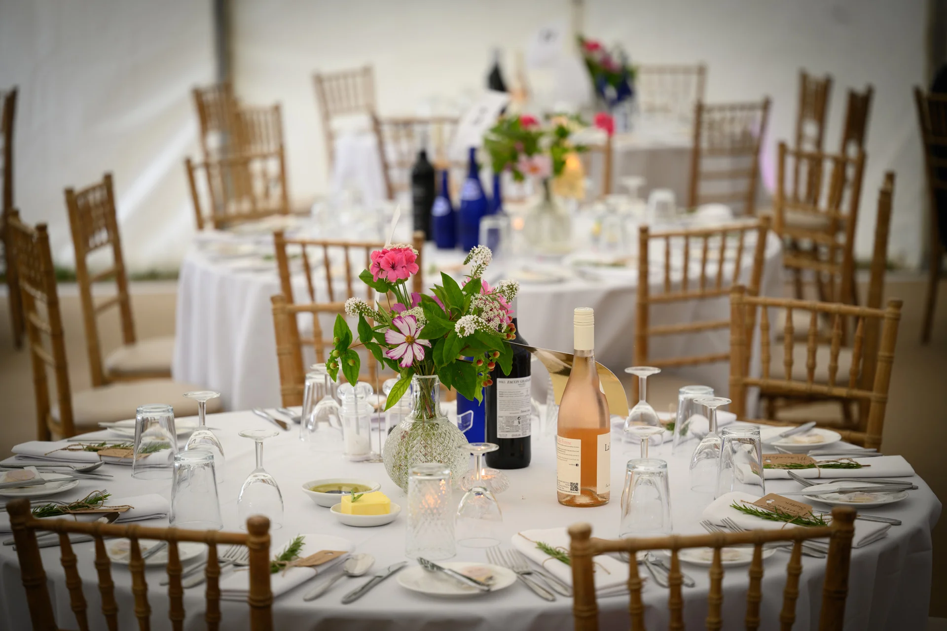A beautifully set round wedding table inside a marquee, featuring white linens, chiavari chairs, delicate pink and white floral centrepieces in bud vases, and wine bottles.