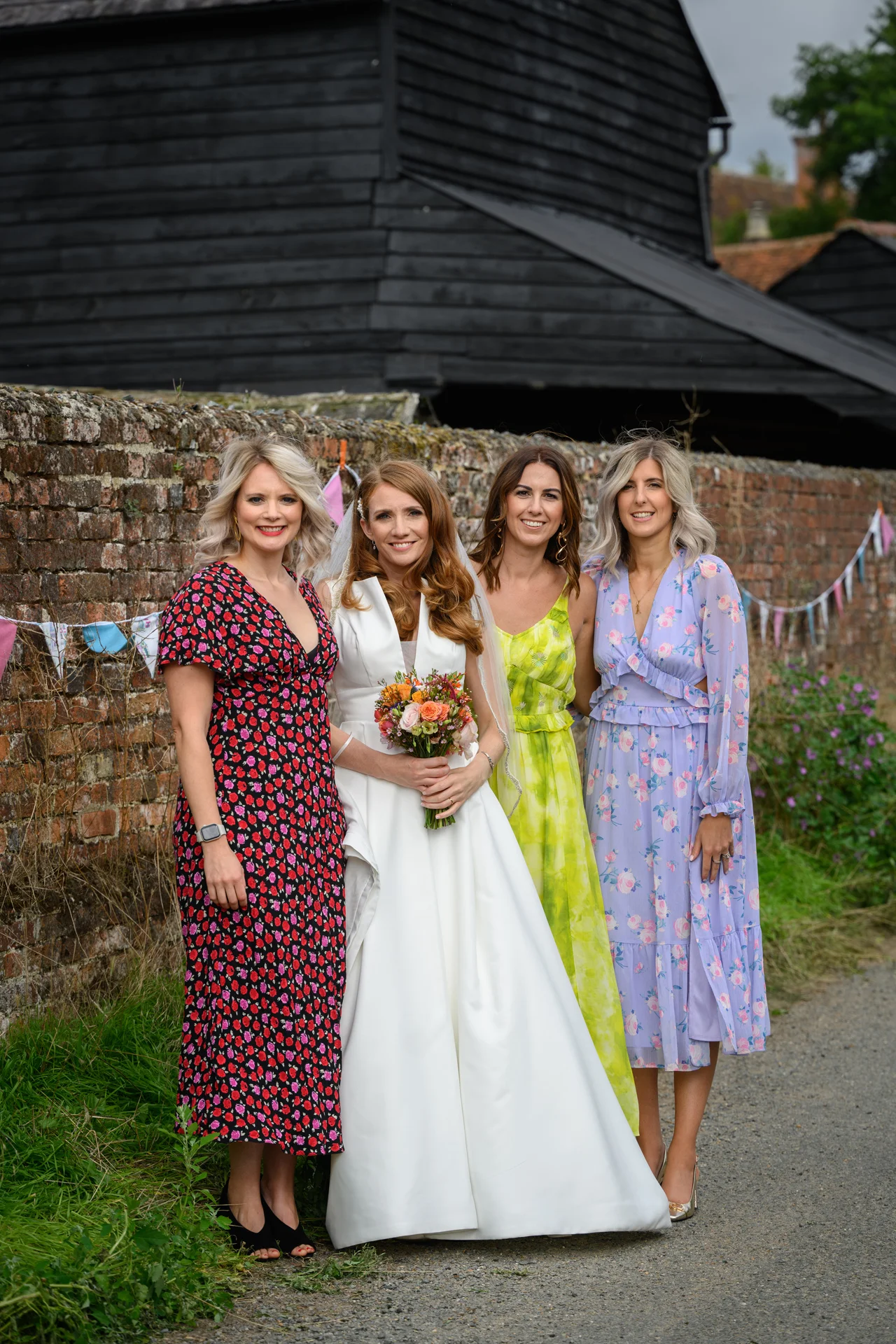 A smiling bride holding a colourful bouquet stands proudly alongside three guests in floral dresses outside a rustic brick and timber barn.