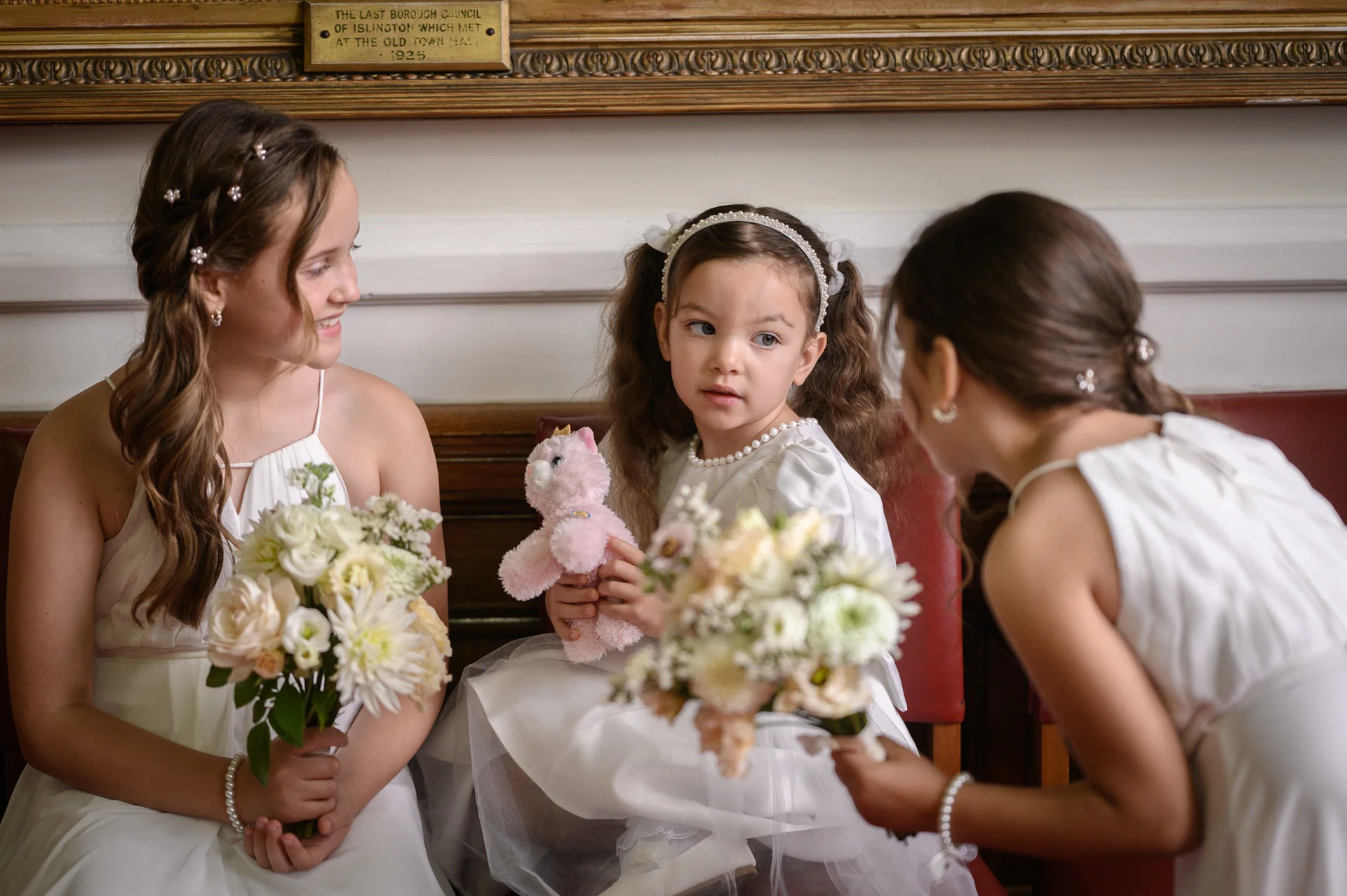 Three young bridesmaids in matching white dresses hold small floral bouquets and a pink toy while interacting together indoors.