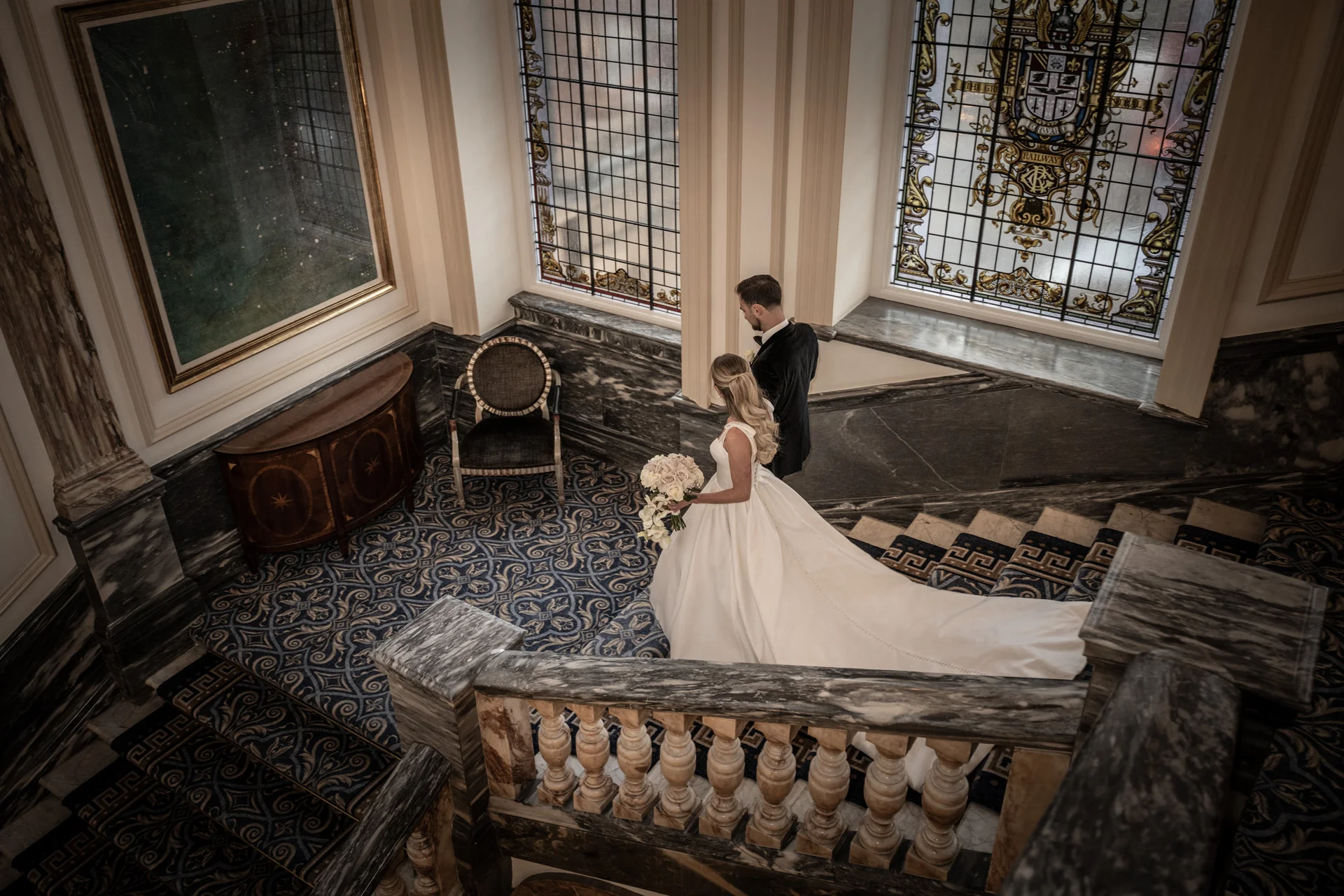 A newlywed couple descending a grand stone staircase inside a luxury hotel ballroom, featuring stained glass windows, ornate marble balustrades, and patterned carpeting.