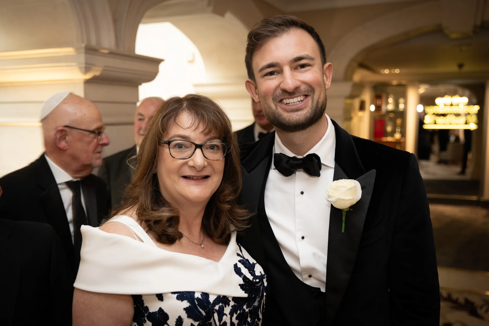 A smiling groom wearing a black tuxedo with a white rose buttonhole poses alongside a female guest inside an elegant venue during the wedding reception.