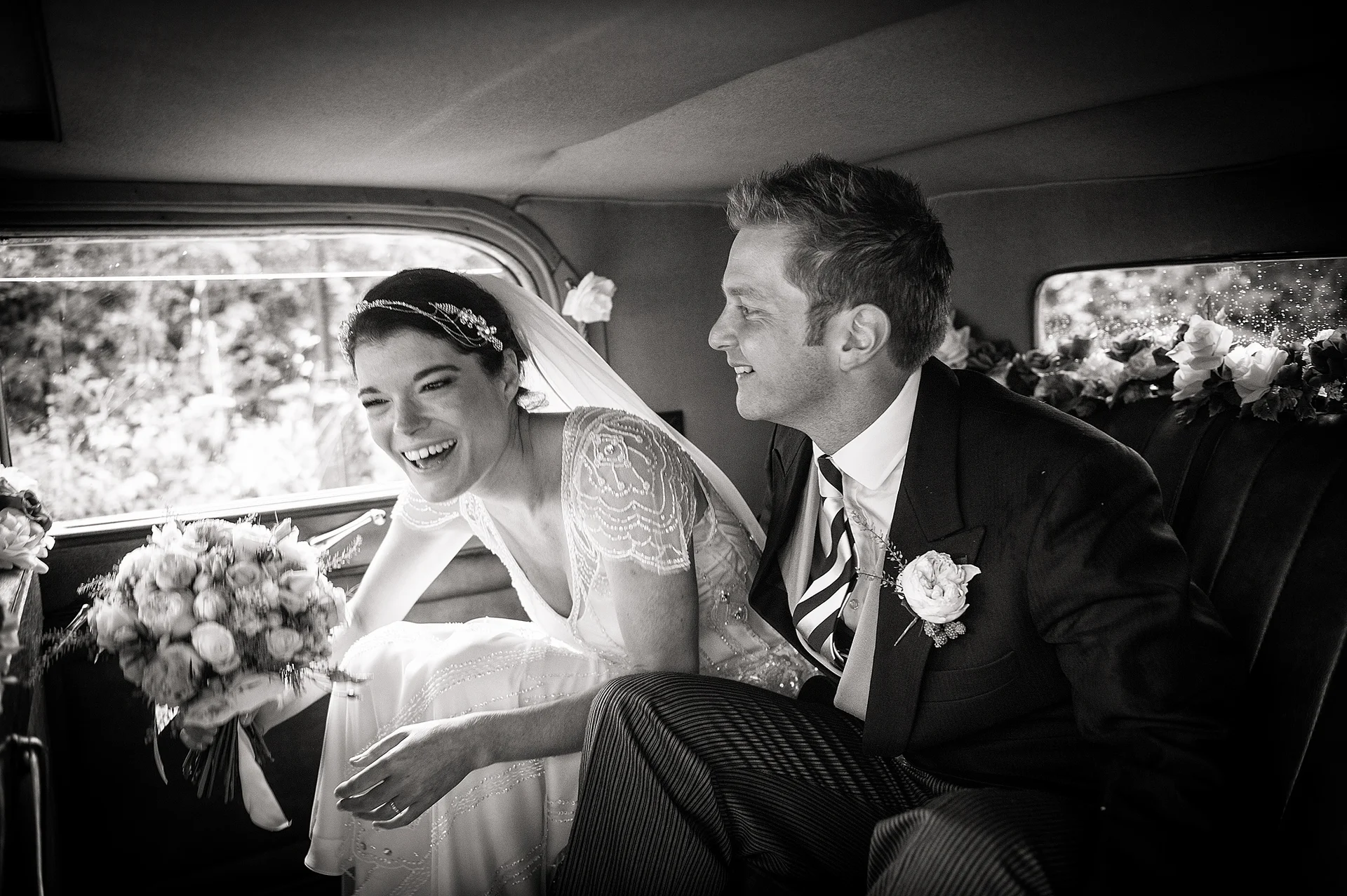 A newlywed couple share a joyful laugh while sitting together in the back of a vintage wedding car, with the bride holding a beautiful bouquet.