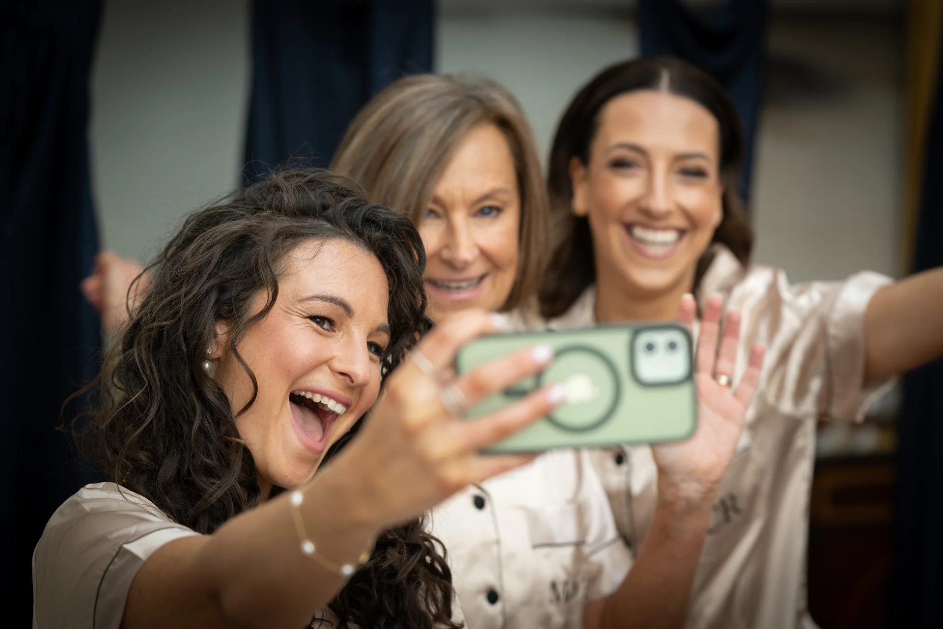 Three smiling women wearing matching silk pyjamas laugh excitedly while taking a selfie together during wedding morning preparations.