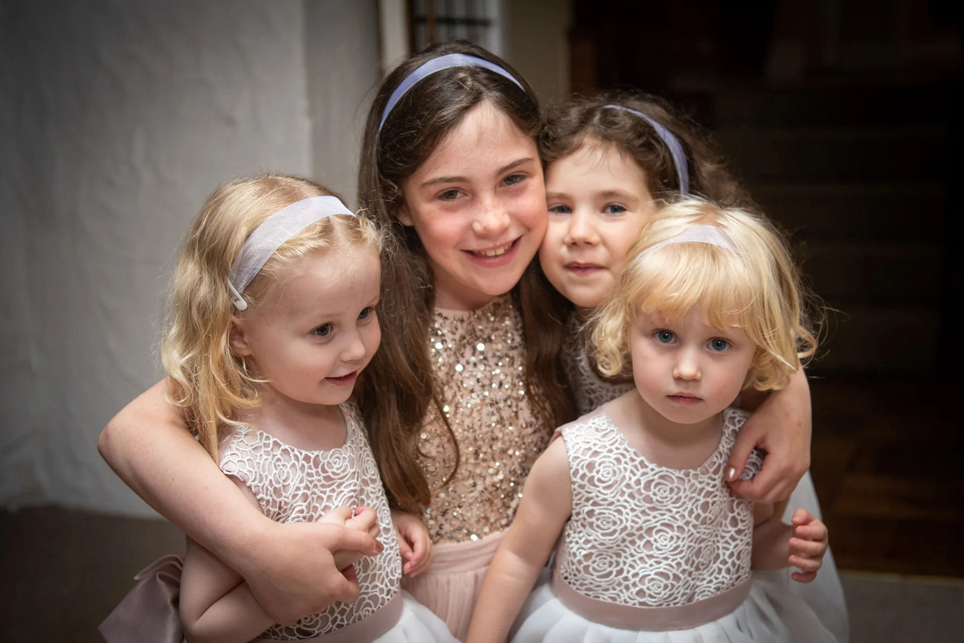 Four young flower girls and junior bridesmaids pose together wearing matching dresses and headbands during a wedding celebration.