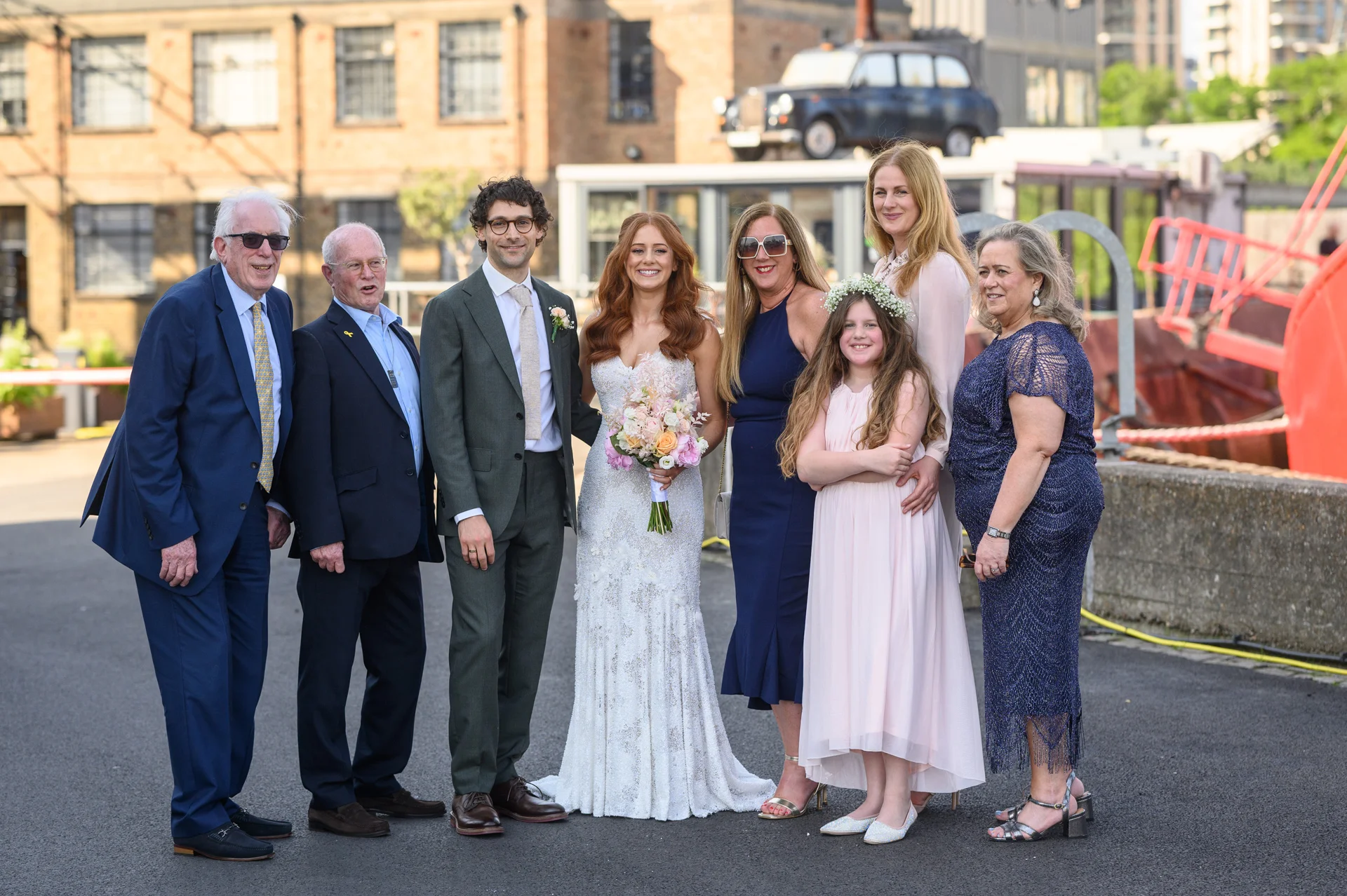 A newlywed couple and their family members pose outdoors in an urban harbour setting for a formal group wedding portrait.