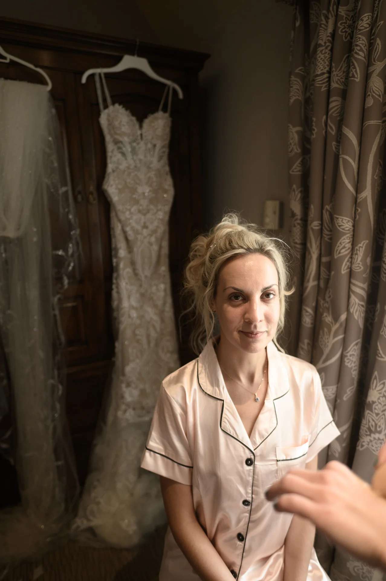 A glowing bride sits comfortably in pink satin pyjamas during morning preparations, with her intricate lace wedding dress hanging in the background.