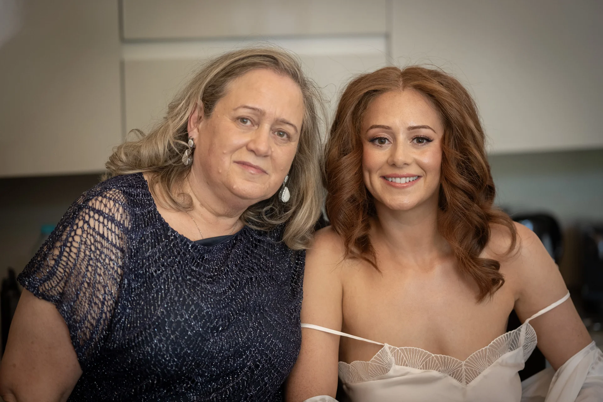 A smiling bride with auburn hair and an off-the-shoulder gown poses beside her mother during morning preparations.