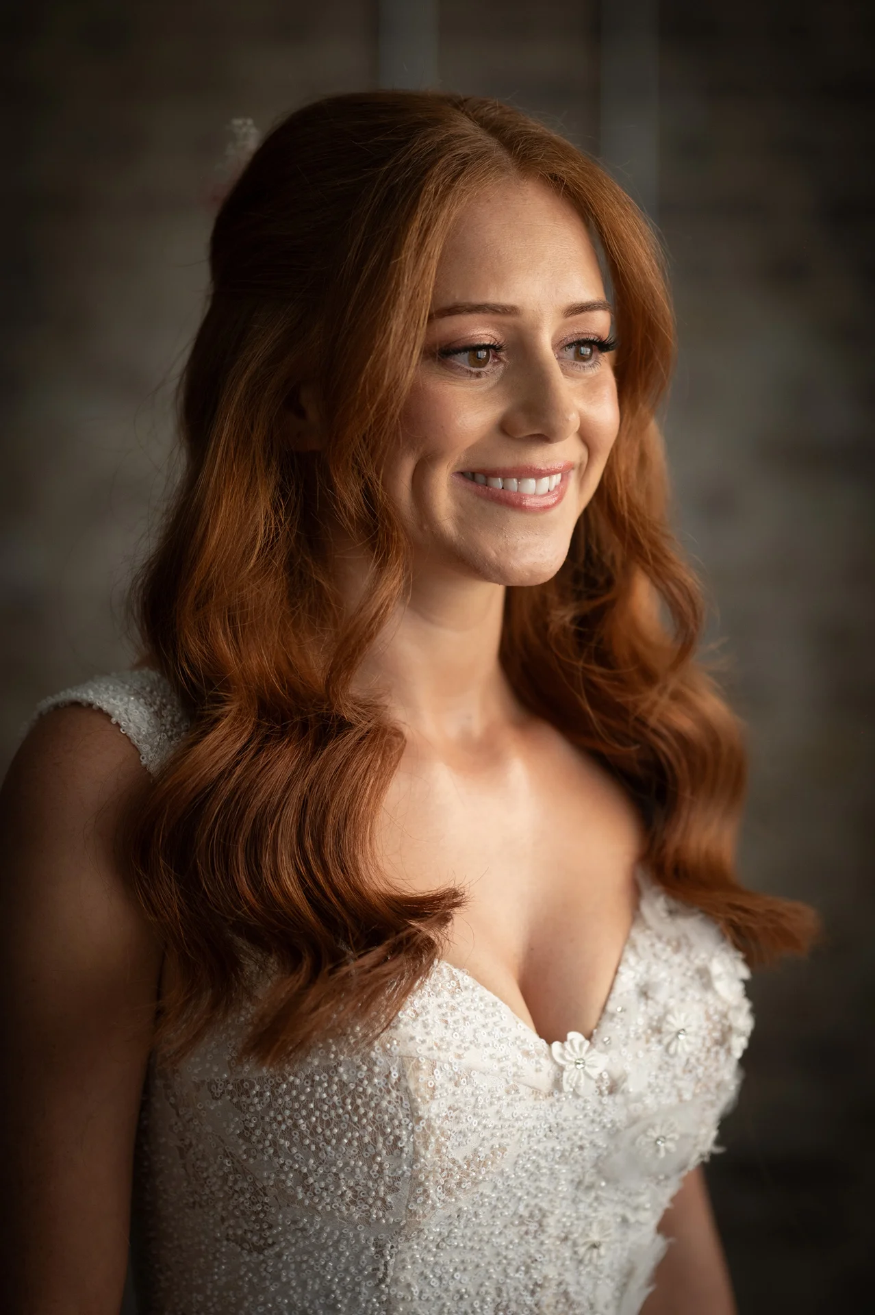 A close-up portrait of a smiling bride with vibrant red hair styled in soft half-up waves, wearing an embellished v-neck wedding dress.
