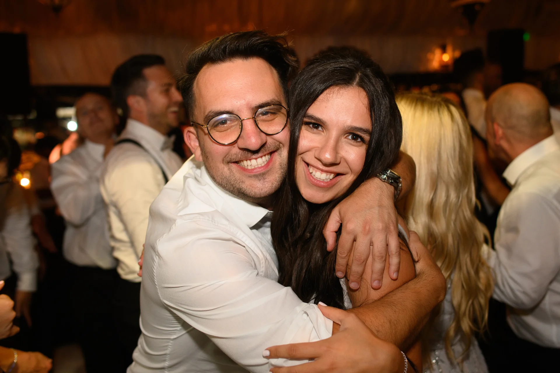 A happy couple smiling and embracing tightly on a lively evening reception dance floor surrounded by wedding guests.