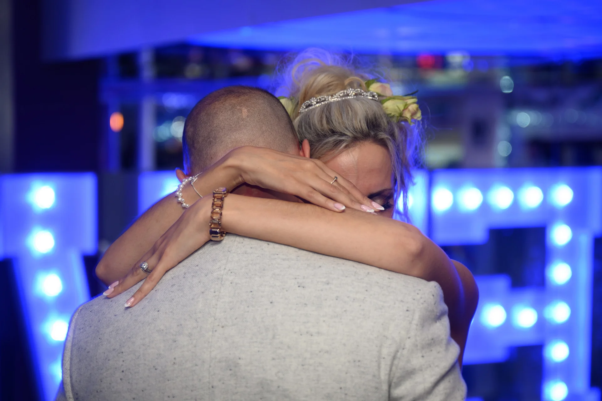 A newlywed couple share a tender embrace during their first dance, illuminated by vibrant blue lighting against a glowing backdrop.