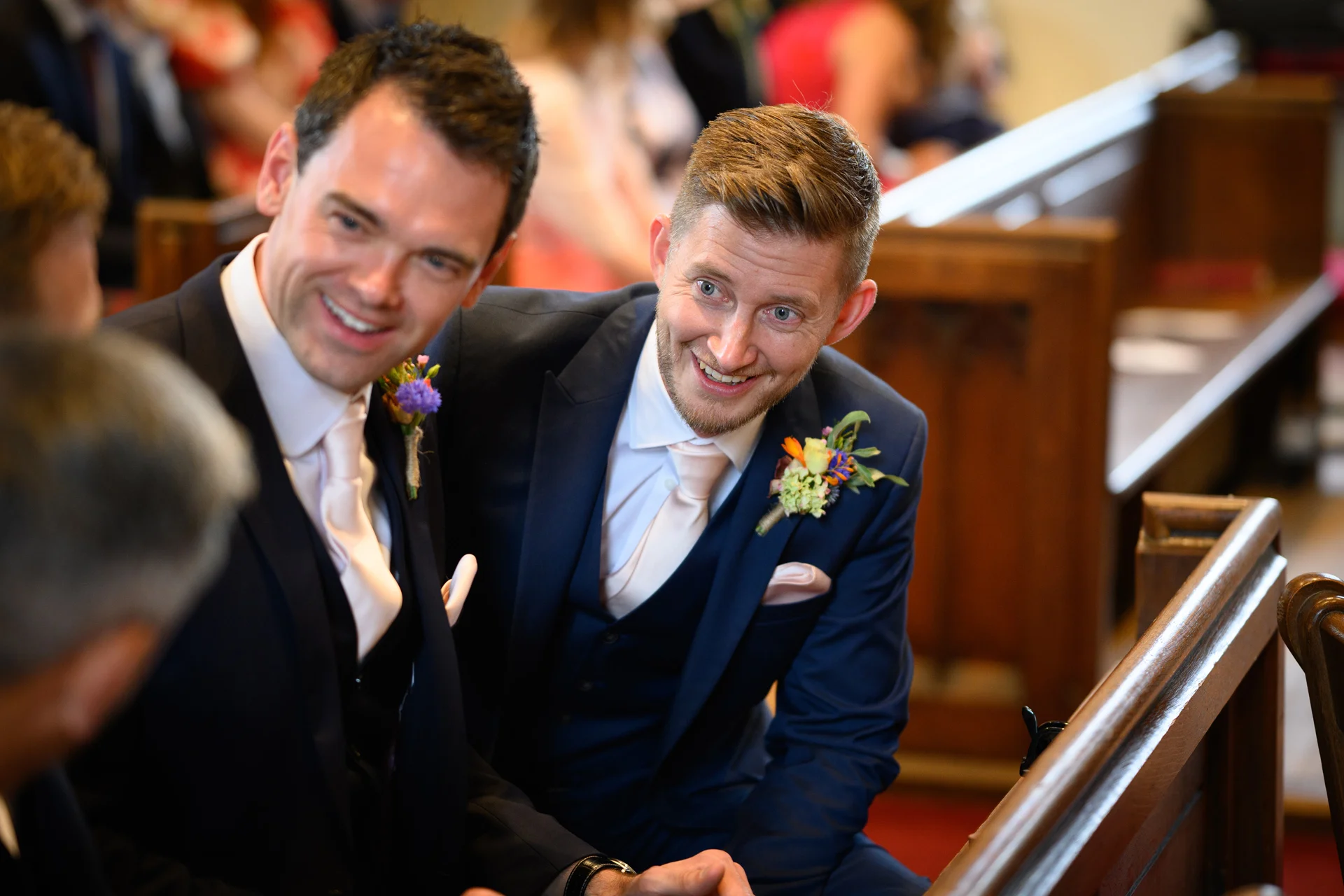 Two grooms in matching navy suits and buttonholes share a happy moment seated in church pews during their ceremony.