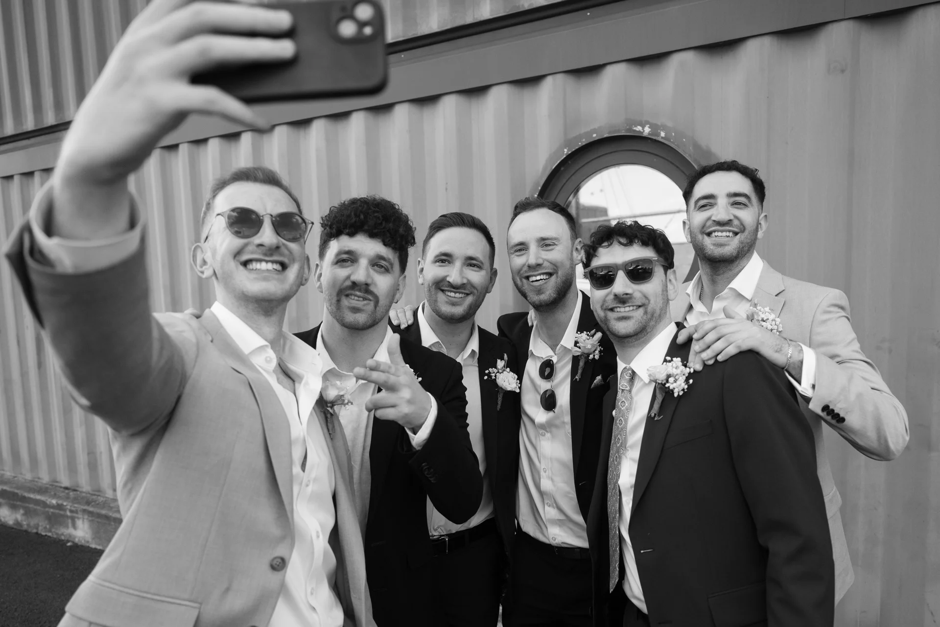 A smiling group of groomsmen take a fun selfie together outside an industrial shipping container venue.