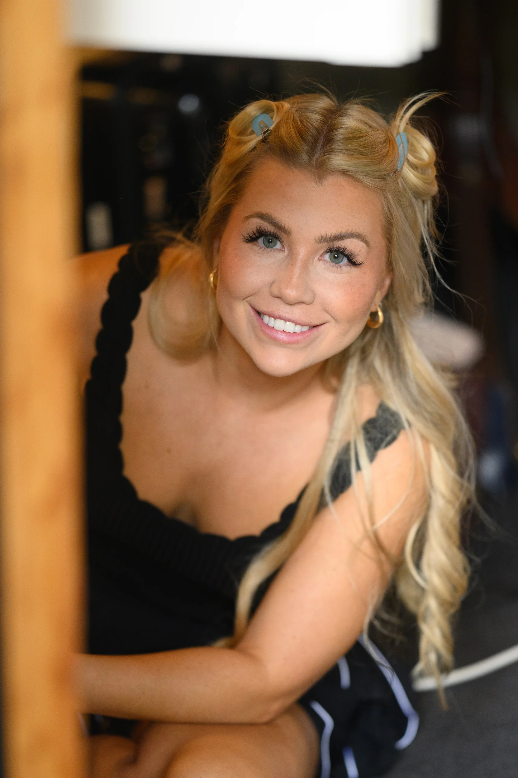 A smiling blonde bride with her hair styled in half-up space buns looks up cheerfully during bridal morning preparations.