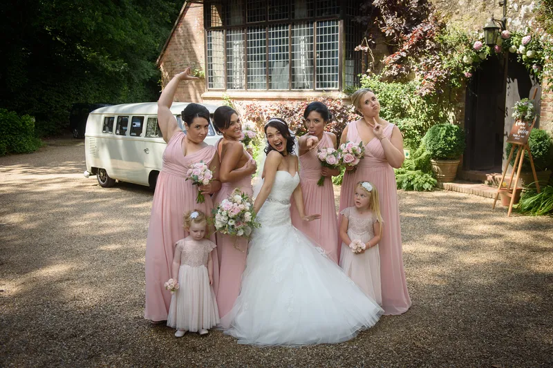 The bride and her playful bridal party in pink dresses pose with humorous expressions outside a historic country house next to a vintage campervan.