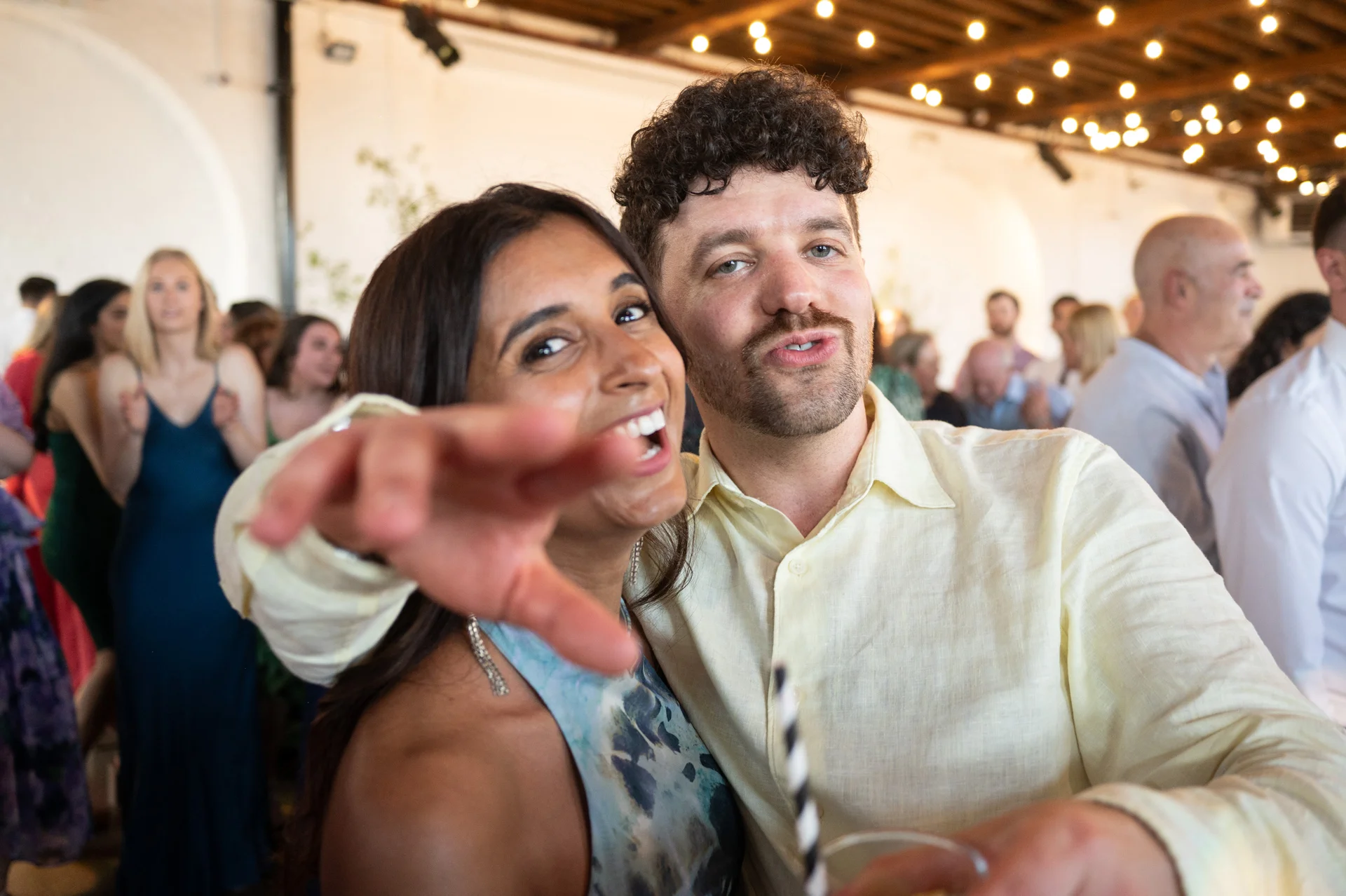 A joyous couple laughing and posing playfully on a lively crowded dance floor illuminated by overhead festoon lights.