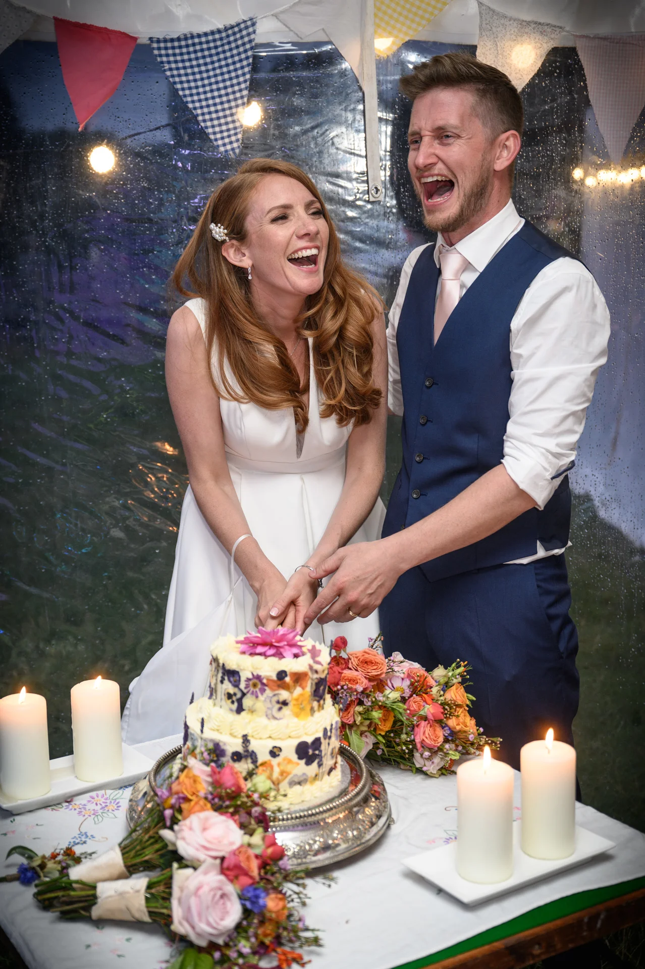 A joyful newlywed couple laugh heartily while cutting their two-tier pressed flower wedding cake inside a candlelit marquee adorned with colourful bunting.